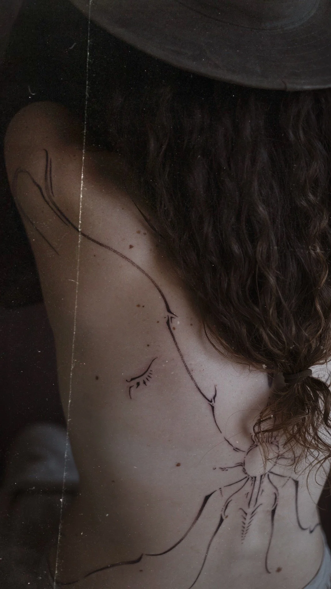 Close-up of a person's back with black ink line art of a bicycle and a single eyelash design drawn on their skin, with a woman having curly brown hair tied back and lying down.