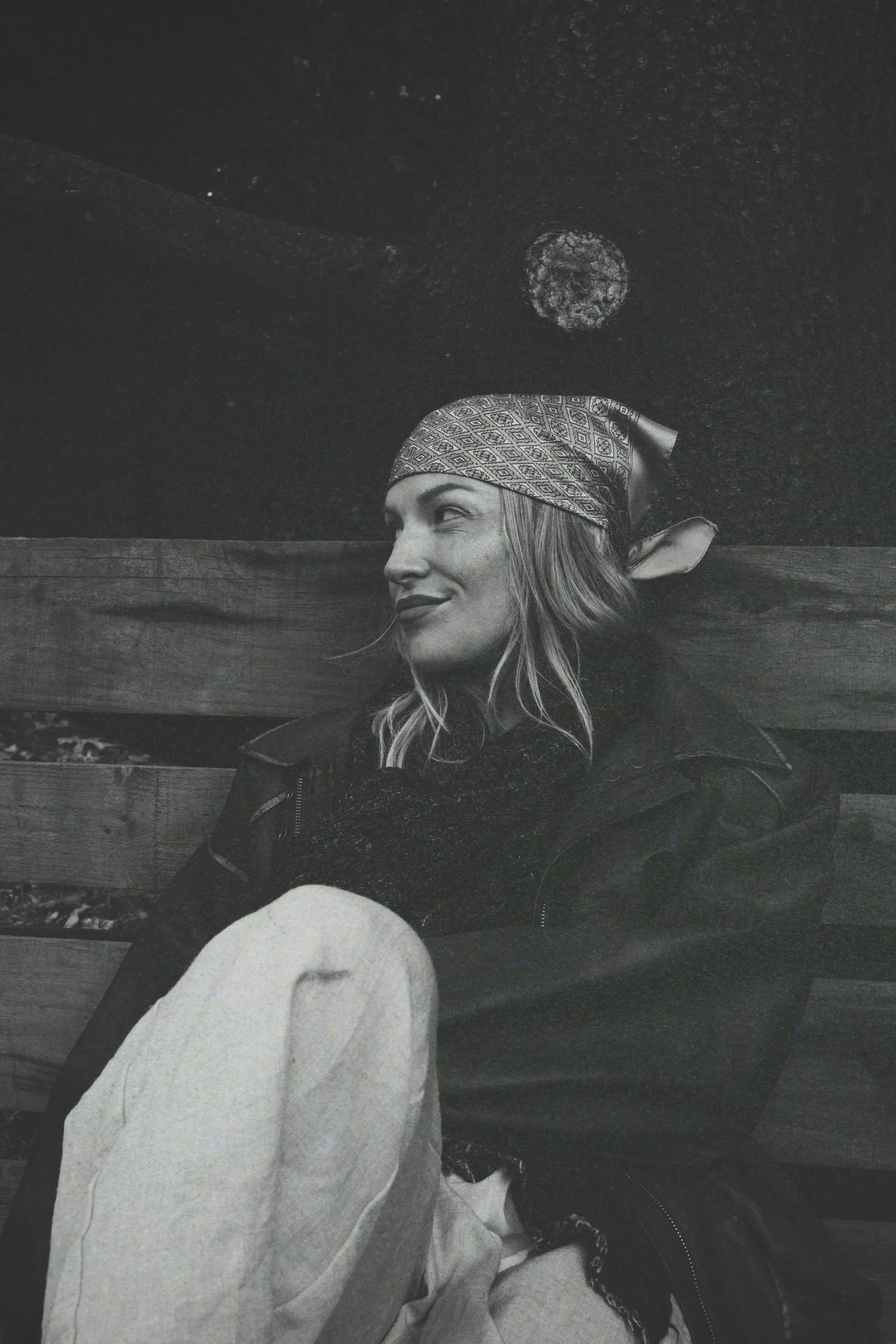 A black and white photo of a woman wearing a bandana, sitting outdoors on a wooden bench with a woman in the background and a starry night sky above.