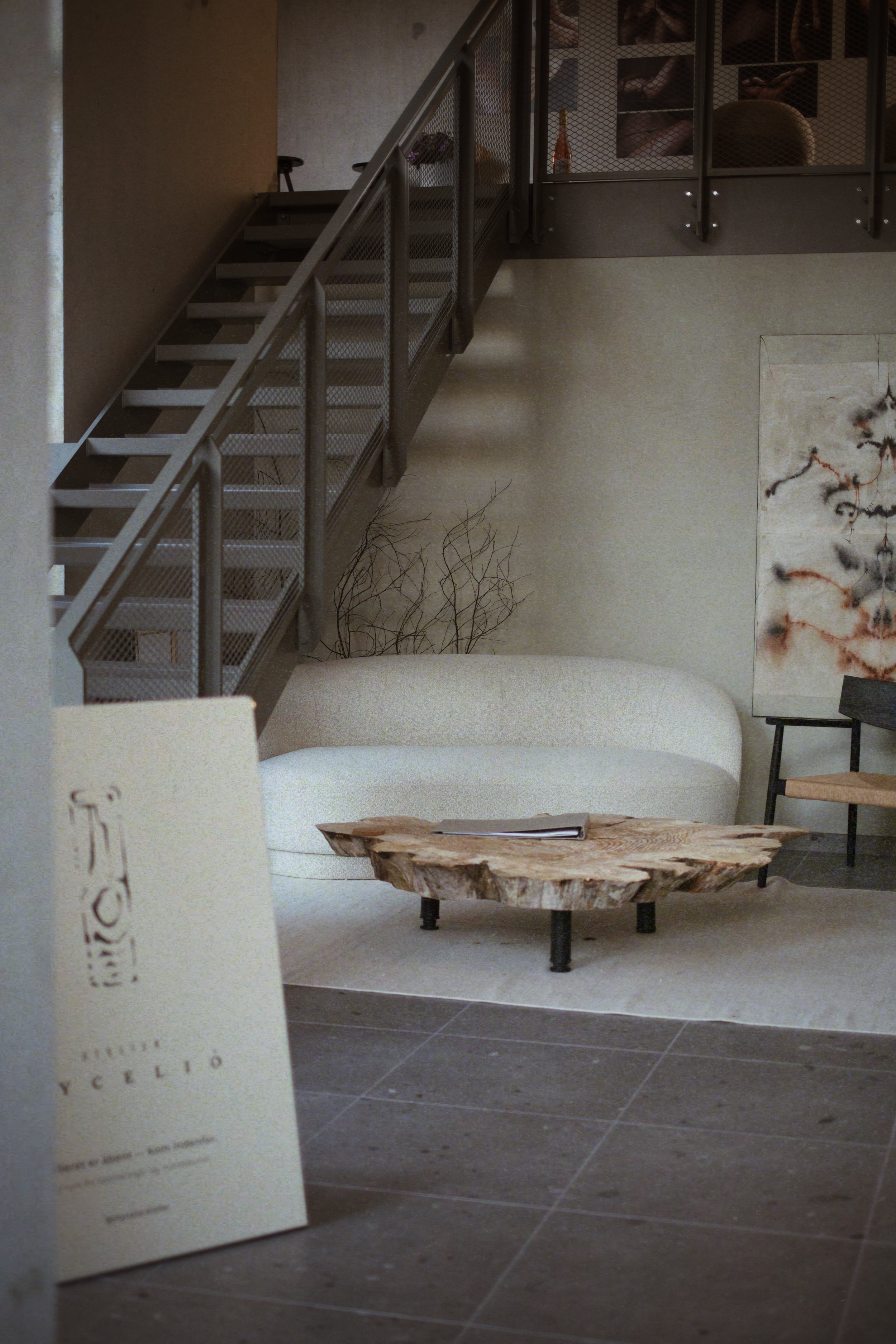 A modern interior living room with a beige curved sofa, a rustic wooden coffee table, and abstract artwork on the wall. There is a staircase with metal railing leading upstairs and a brochure or menu on the floor.