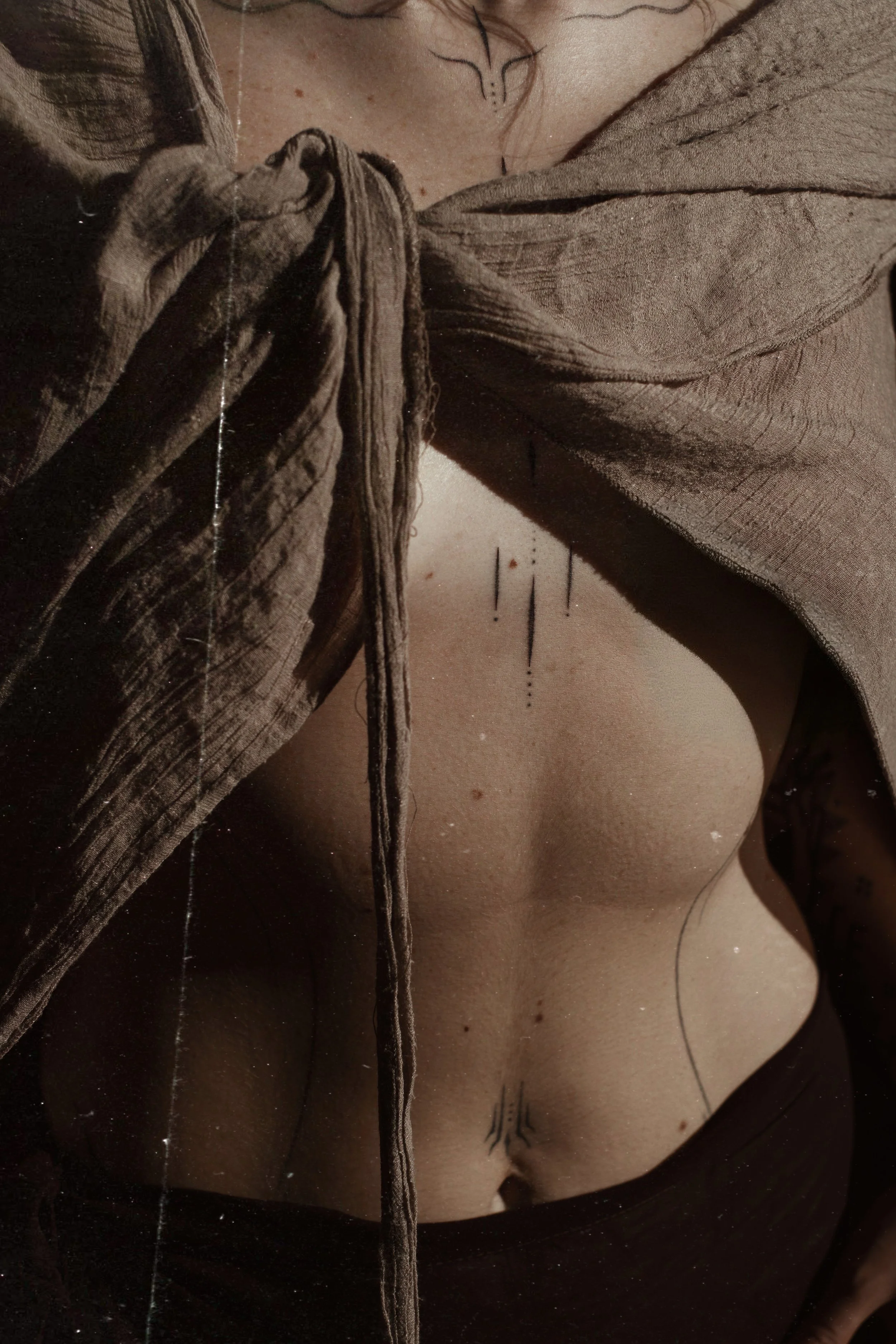 Close-up of a woman's upper torso, showing her bare chest with various small tattoos and a brown, textured, tied shirt partially covering her shoulders.