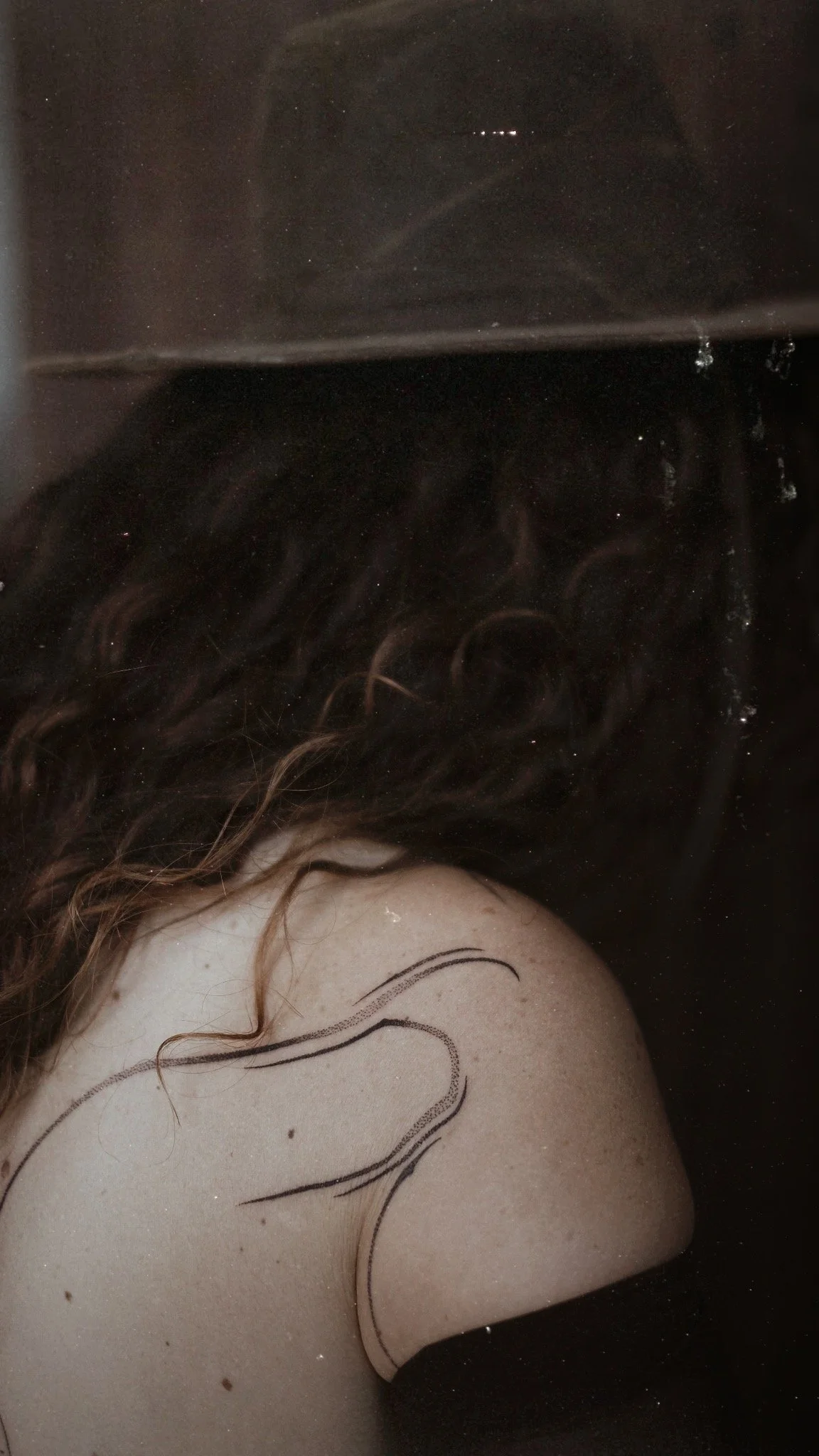 A person with curly hair resting their head on a surface, with a black painted line on their shoulder.