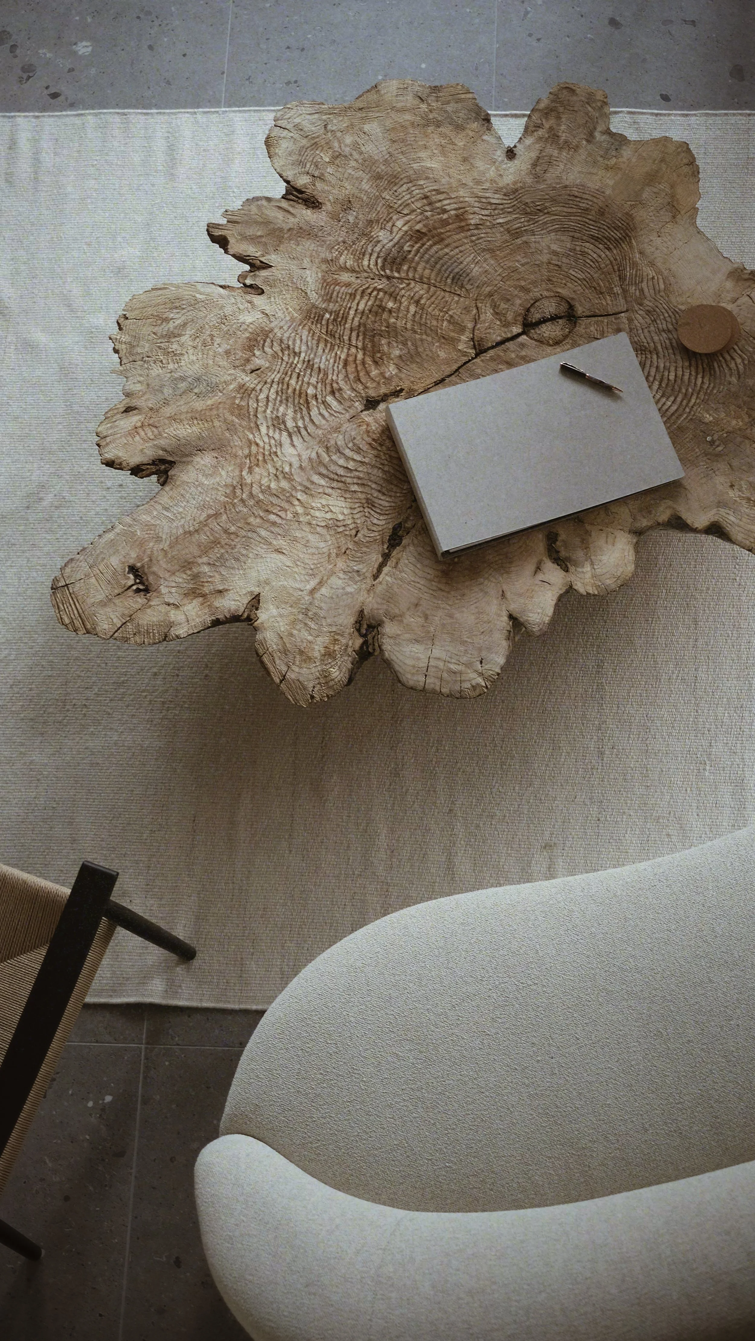 A rustic wooden table with visible wood grain and knots, placed on a beige rug. On the table, there is a closed notebook, a small metallic pen, and a single round object. Partially visible around the table are a beige upholstered chair with curved armrests and a part of a wooden chair with fabric seating.