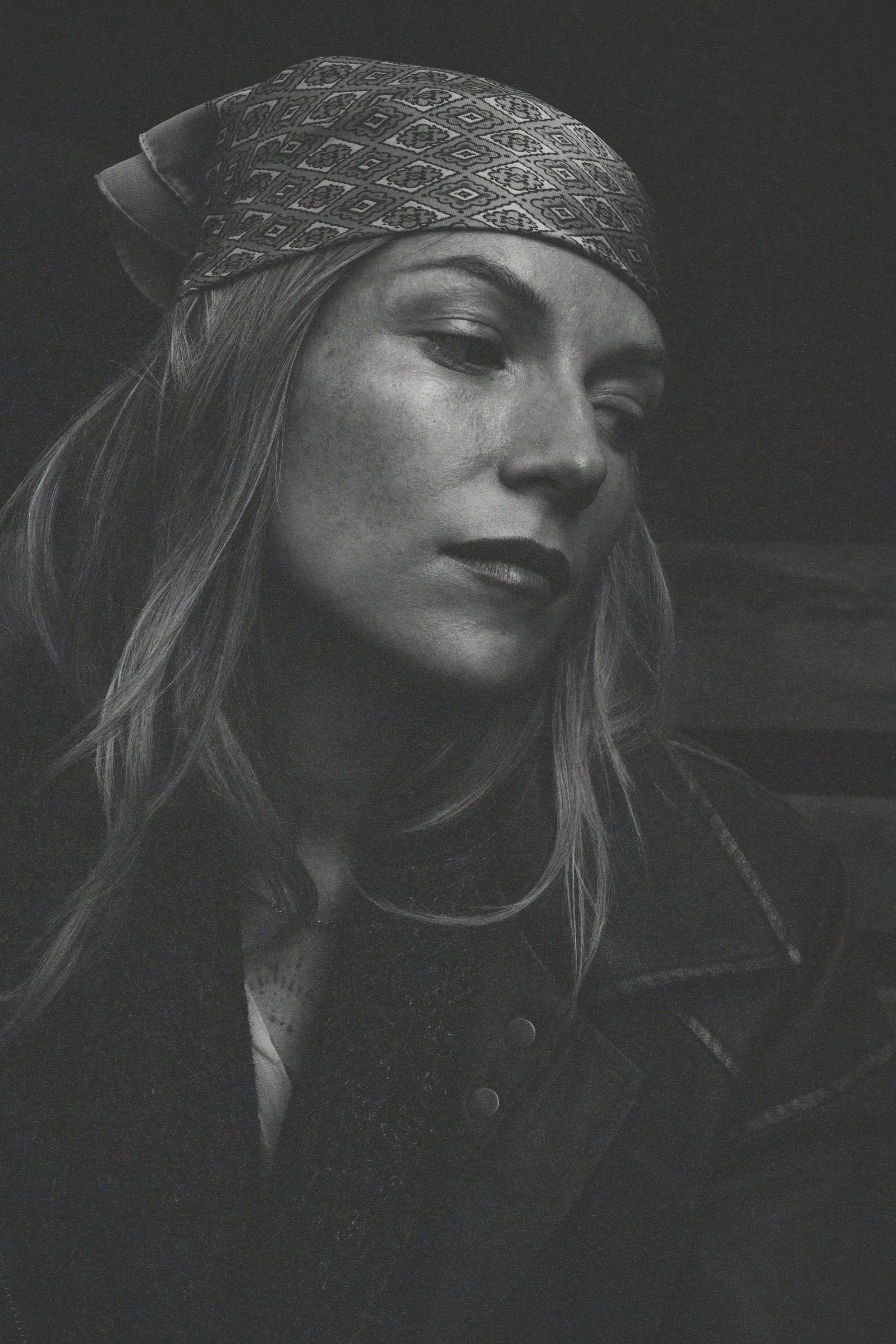 Black and white portrait of a woman wearing a patterned bandana, with long hair, looking slightly downward, with freckles on her face.