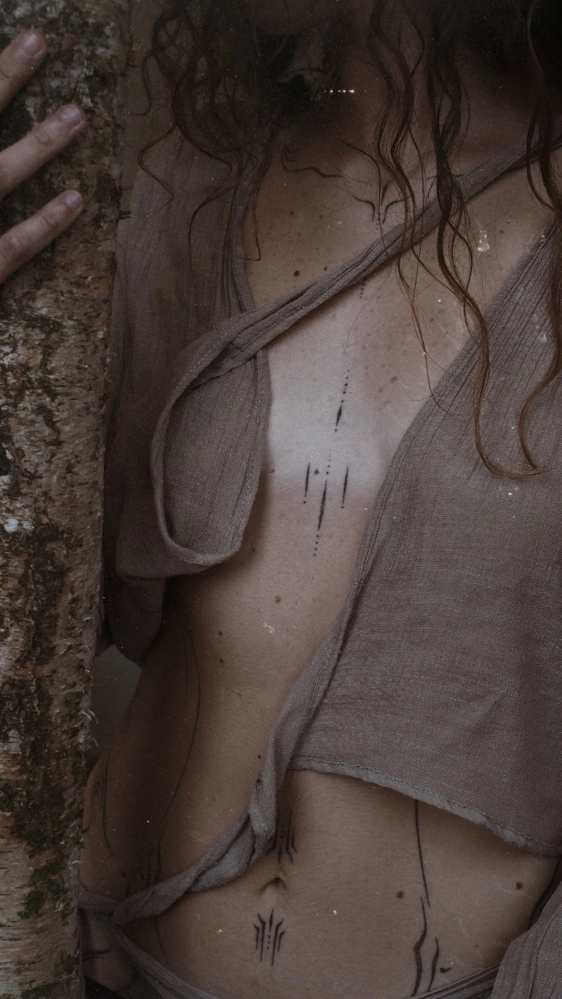 A woman is holding a tree trunk close to her face. She has long curly hair, and is wearing a brown top. The photo captures her chest and the top of her stomach, with some tattoos visible on her skin.