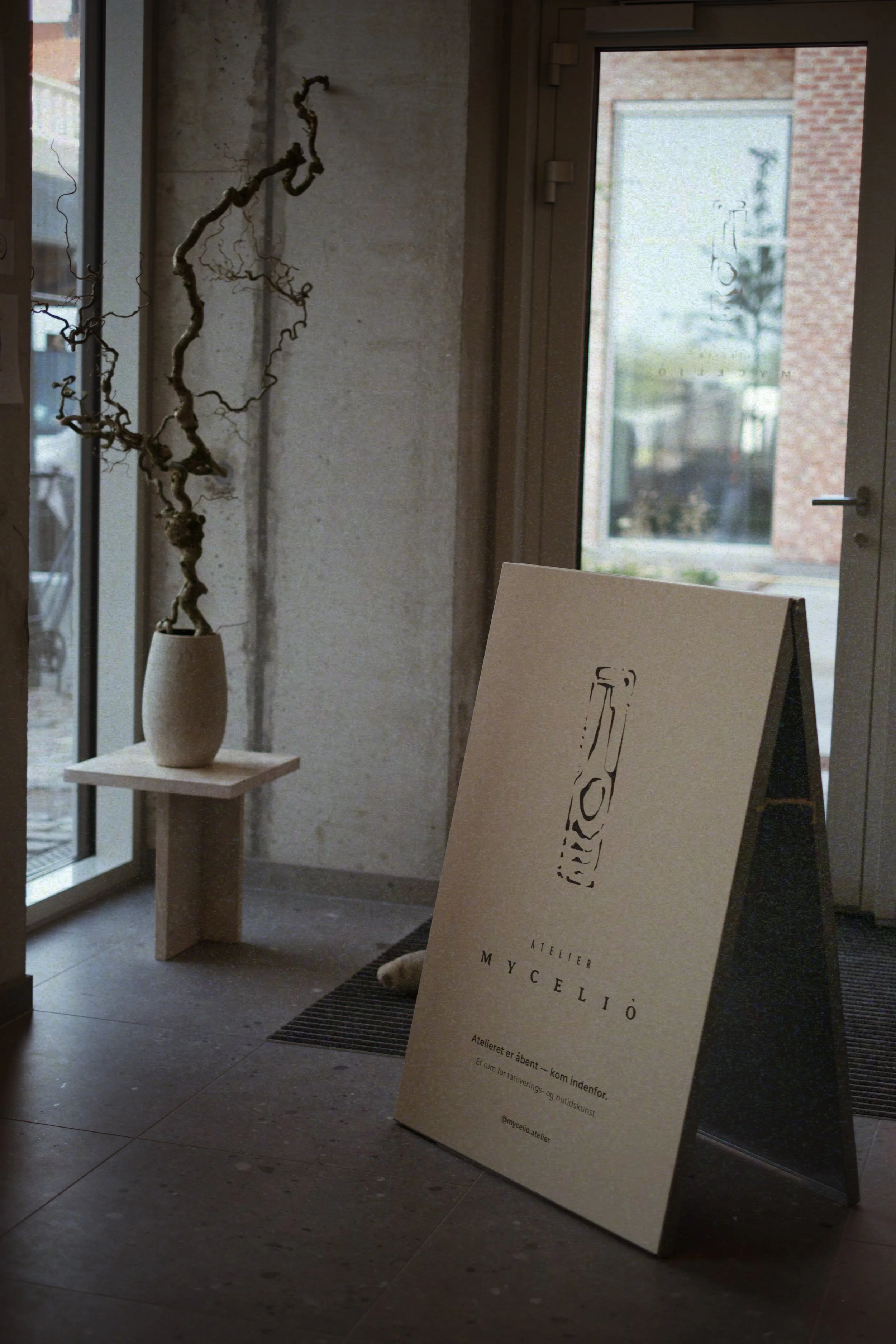 Indoor scene with a large beige signboard on the floor displaying the logo and name 'Atelier Mycéliô', next to a small table holding a vase with twisted branches inside. The background features a glass door and window showing an outdoor view.