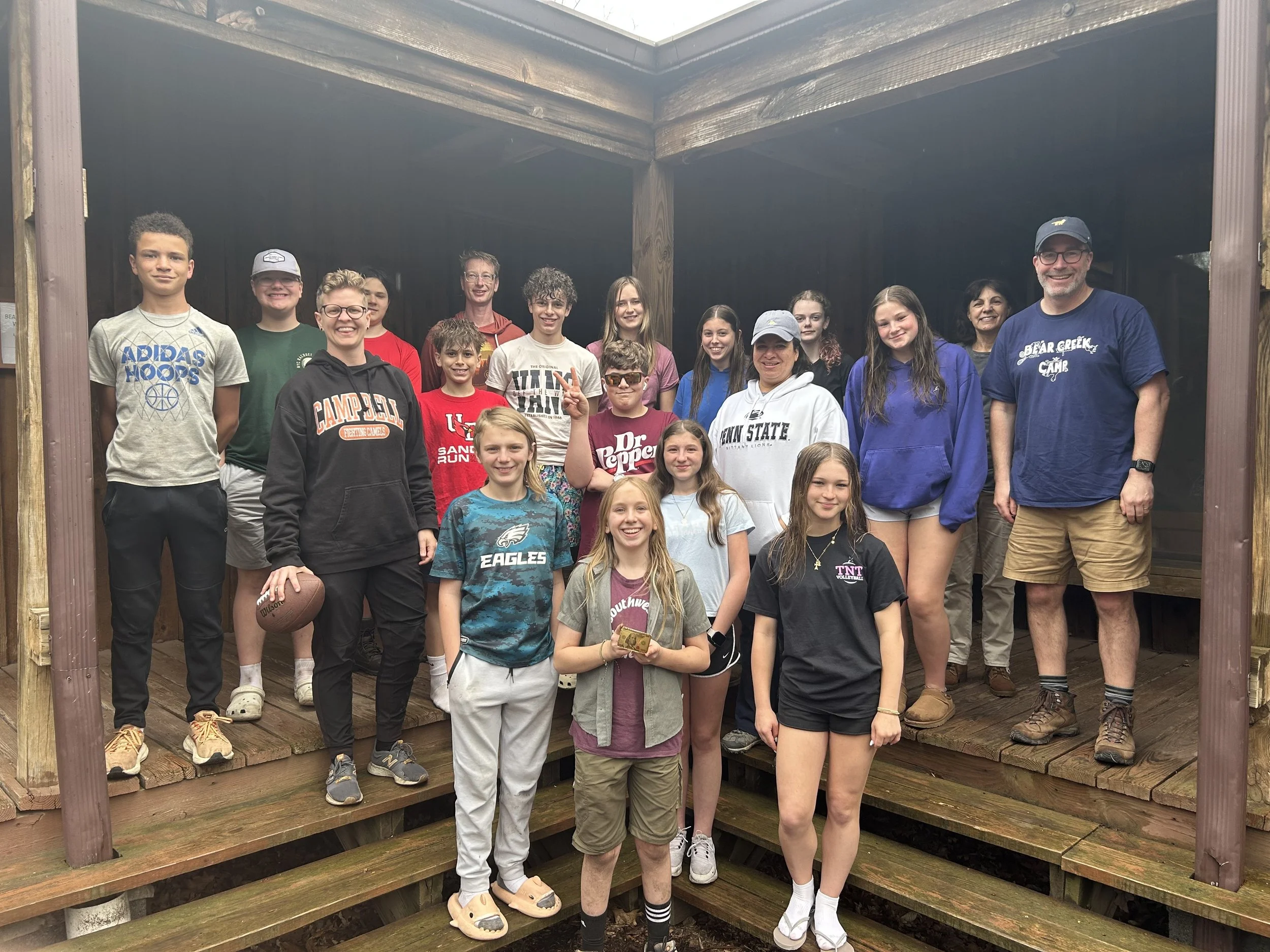 Group photo of children and adults standing on a wooden outdoor stage or platform under a covered shelter at a camp, smiling for the camera.