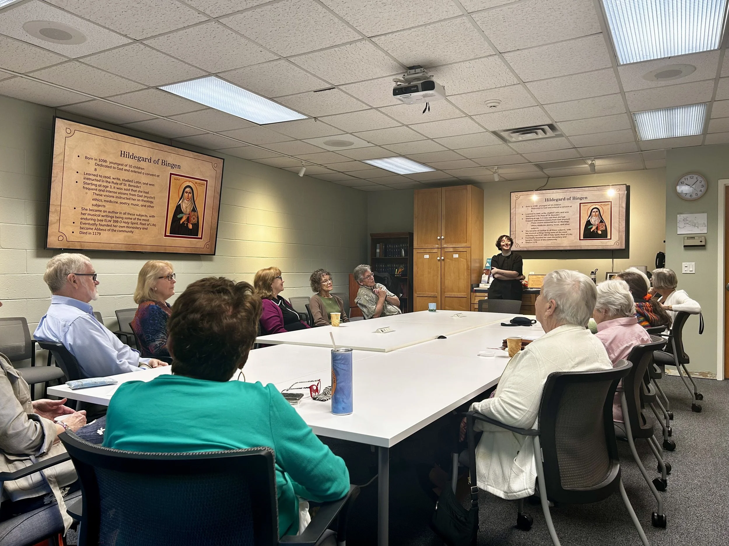 A woman standing and speaking to a group of older adults in a classroom setting, with two large screens displaying information about Hildegard of Bingen on the wall behind her.