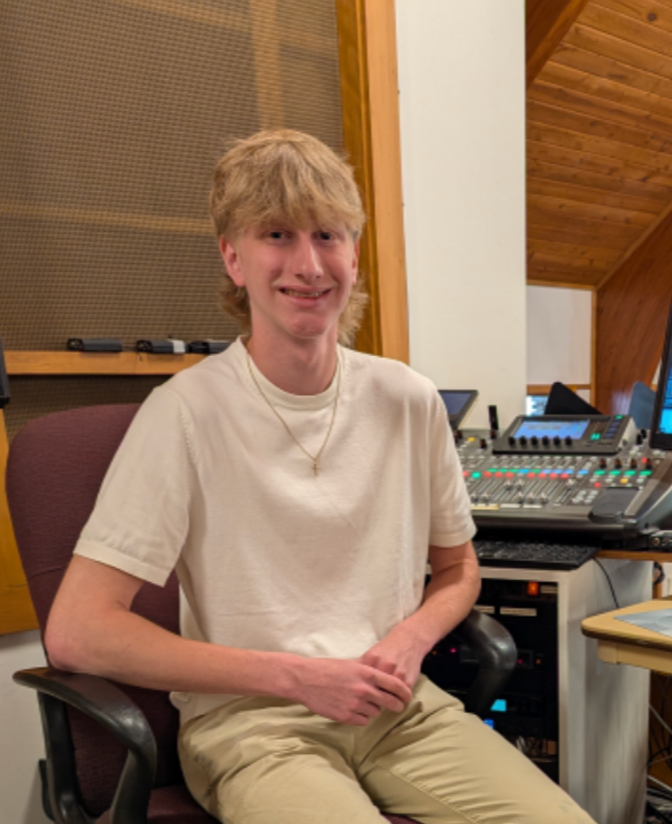 A young man with blonde hair sitting in a recording studio, smiling, wearing a white t-shirt and beige pants, with audio equipment and a computer in the background.