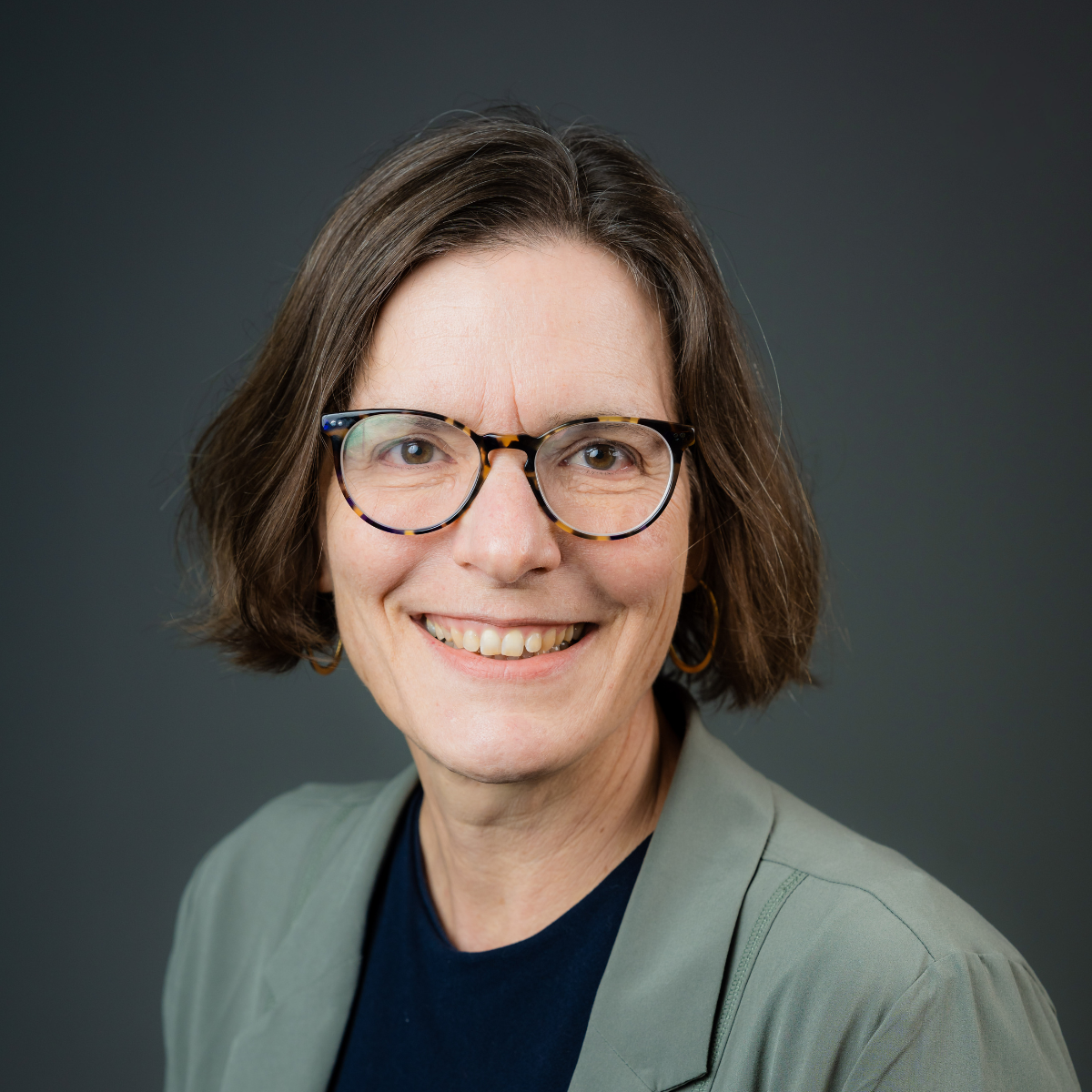 A middle-aged woman with short brown hair, wearing tortoiseshell glasses, gold hoop earrings, a dark blue top, and a light gray blazer, smiling against a dark gray background.