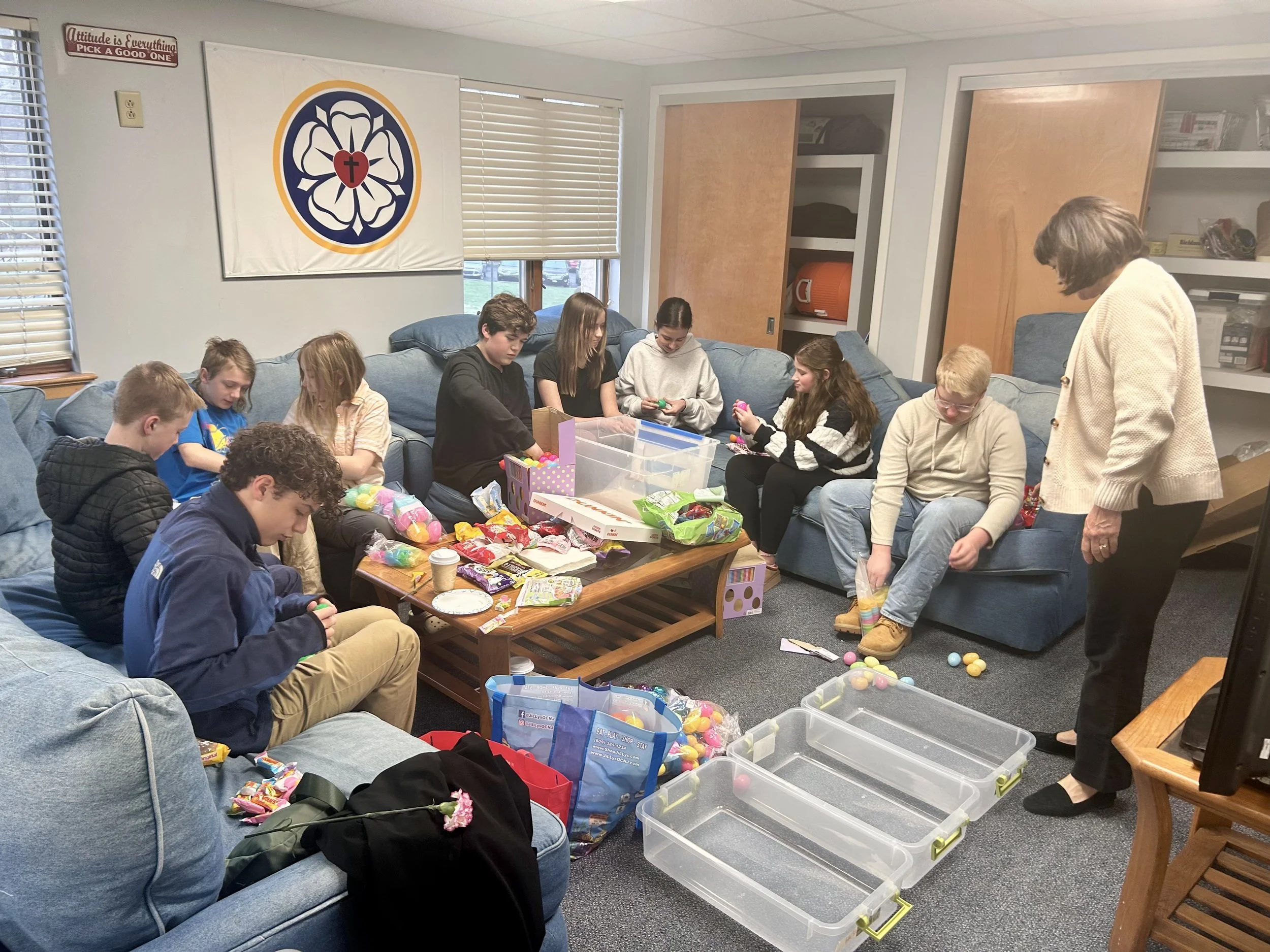 Group of children and adults sitting on couches and sorting colorful plastic eggs and candies in a room decorated with a flag featuring a cross and a flower symbol.