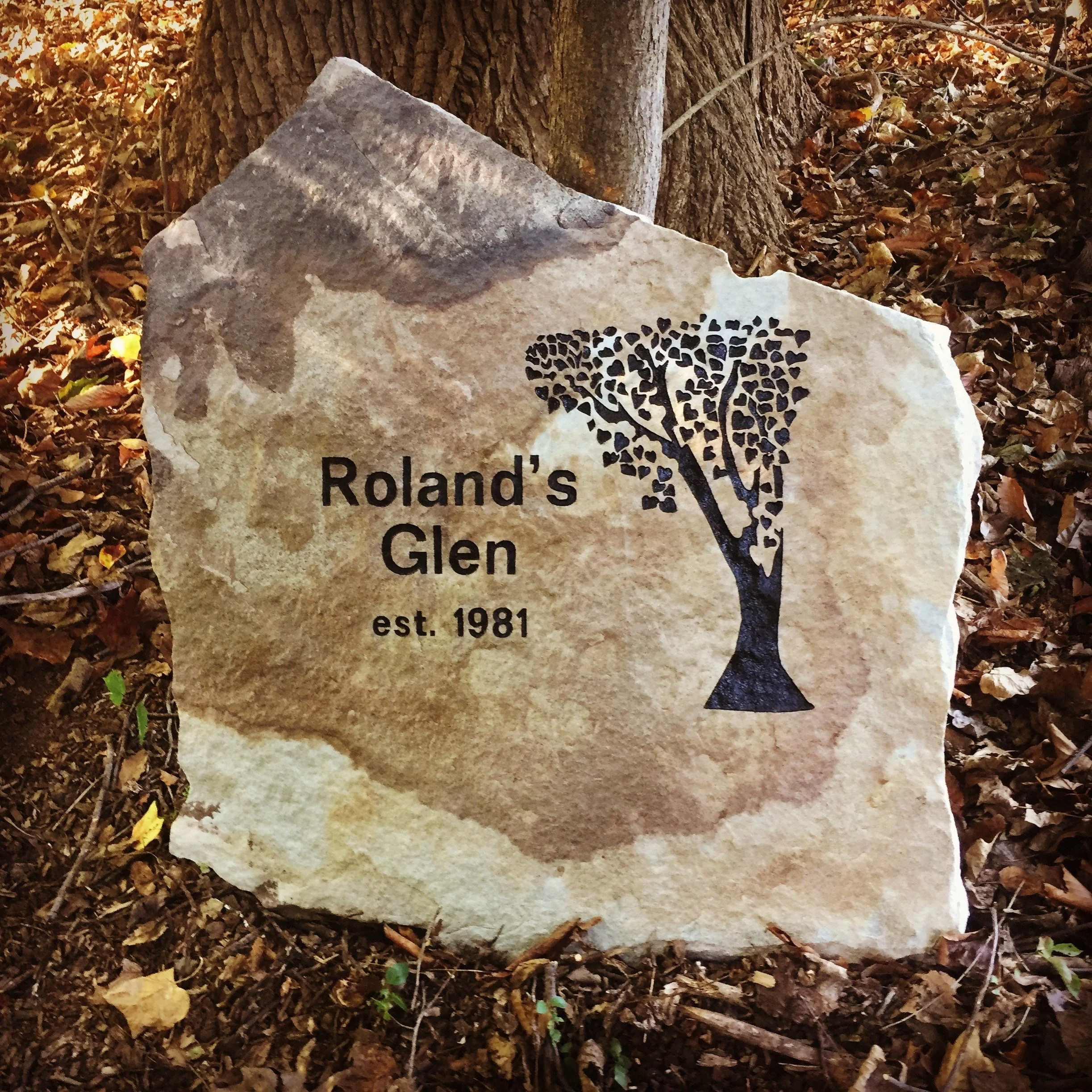 A stone marker with a black tree graphic and the text 'Roland's Glen est. 1981', situated among fallen leaves near a tree trunk.