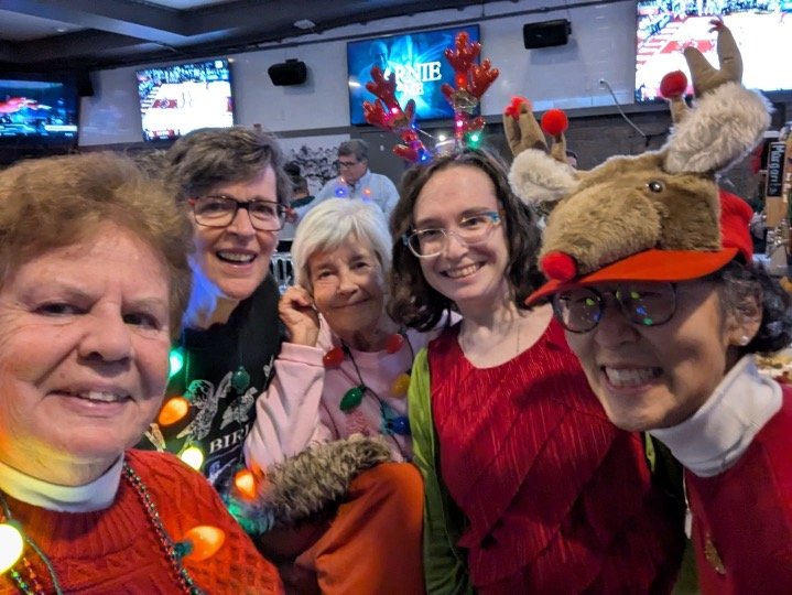 Five friends pose for a festive Christmas selfie in a bar, wearing holiday sweaters, reindeer antlers, and a plush reindeer hat.