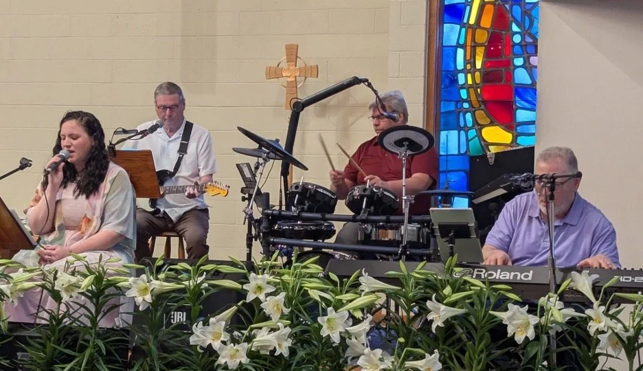 A church band performing on stage with a young woman singing, three men playing instruments including guitar, drums, and keyboard. There are white lilies in front and a colorful stained glass window in the background.