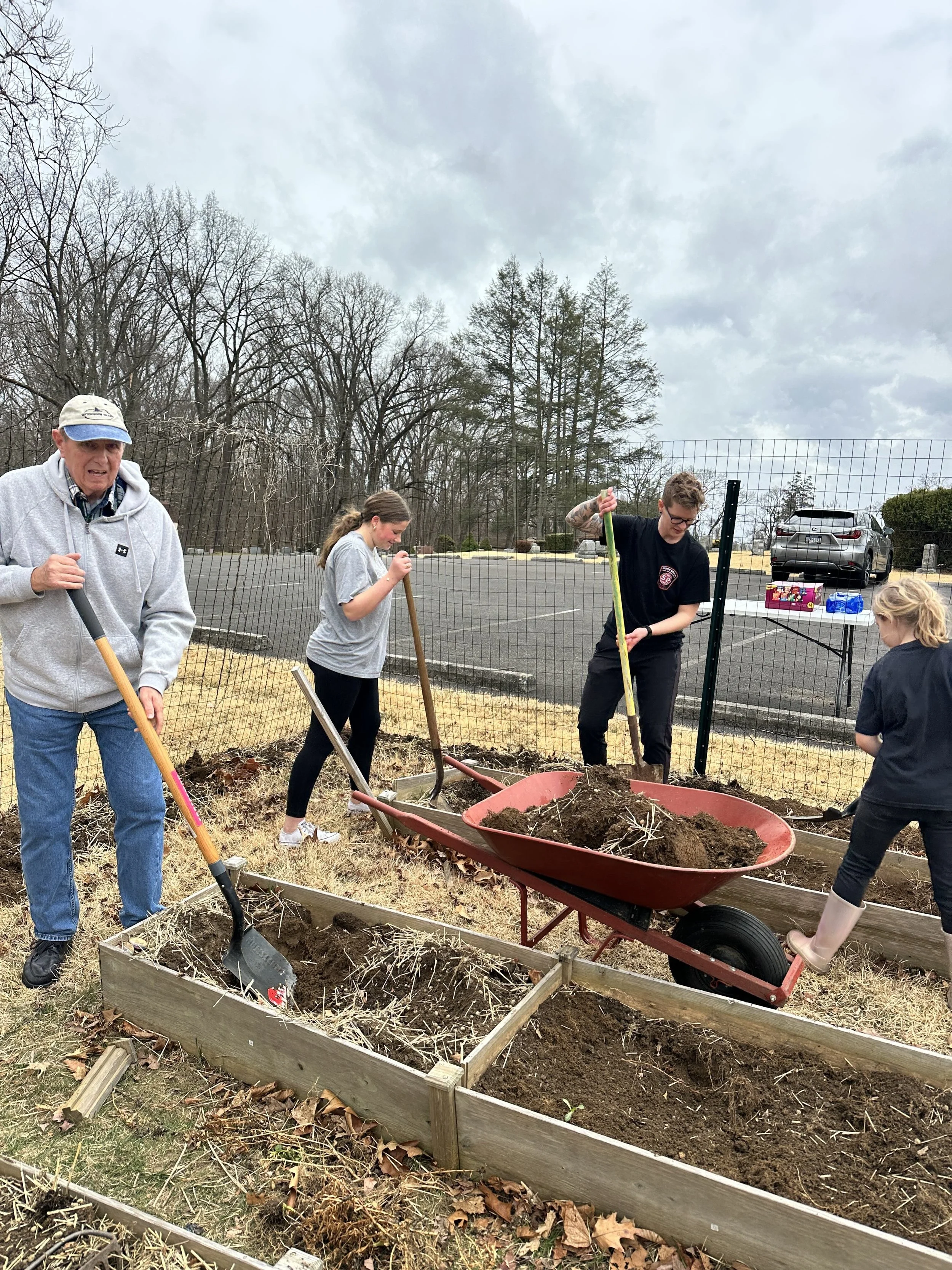 A group of five people, including an elderly man and young girls, working together to dig and prepare garden beds with shovels and a wheelbarrow in an outdoor area.