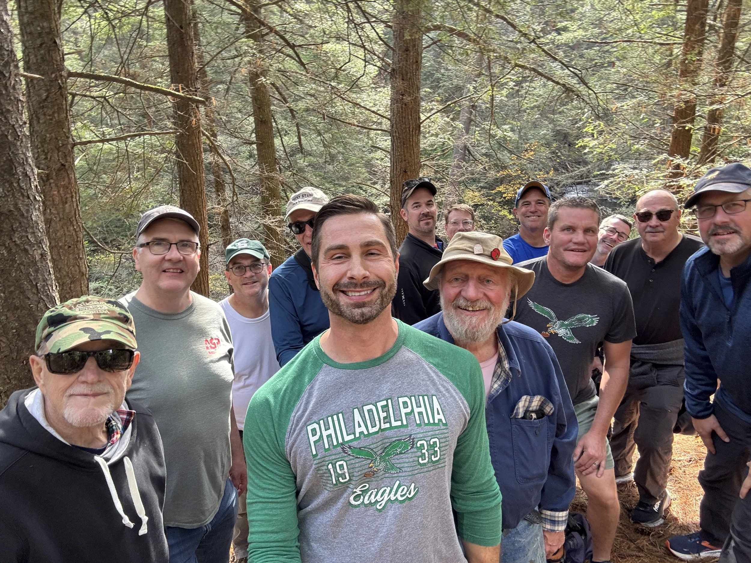 A group of men standing together in a forest, smiling for a photo.