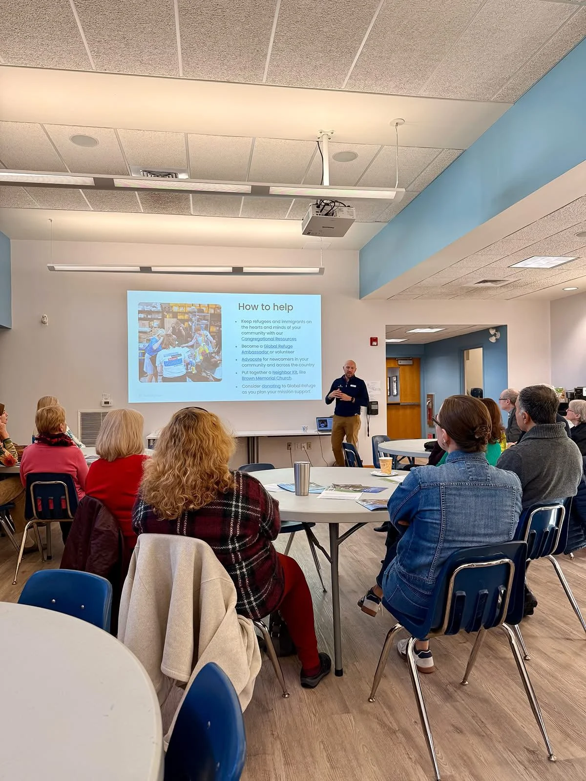 A man speaking at a presentation to a seated group in a room with a large screen displaying a PowerPoint slide titled 'How to help', with bullet points on helping refugees and immigrants.
