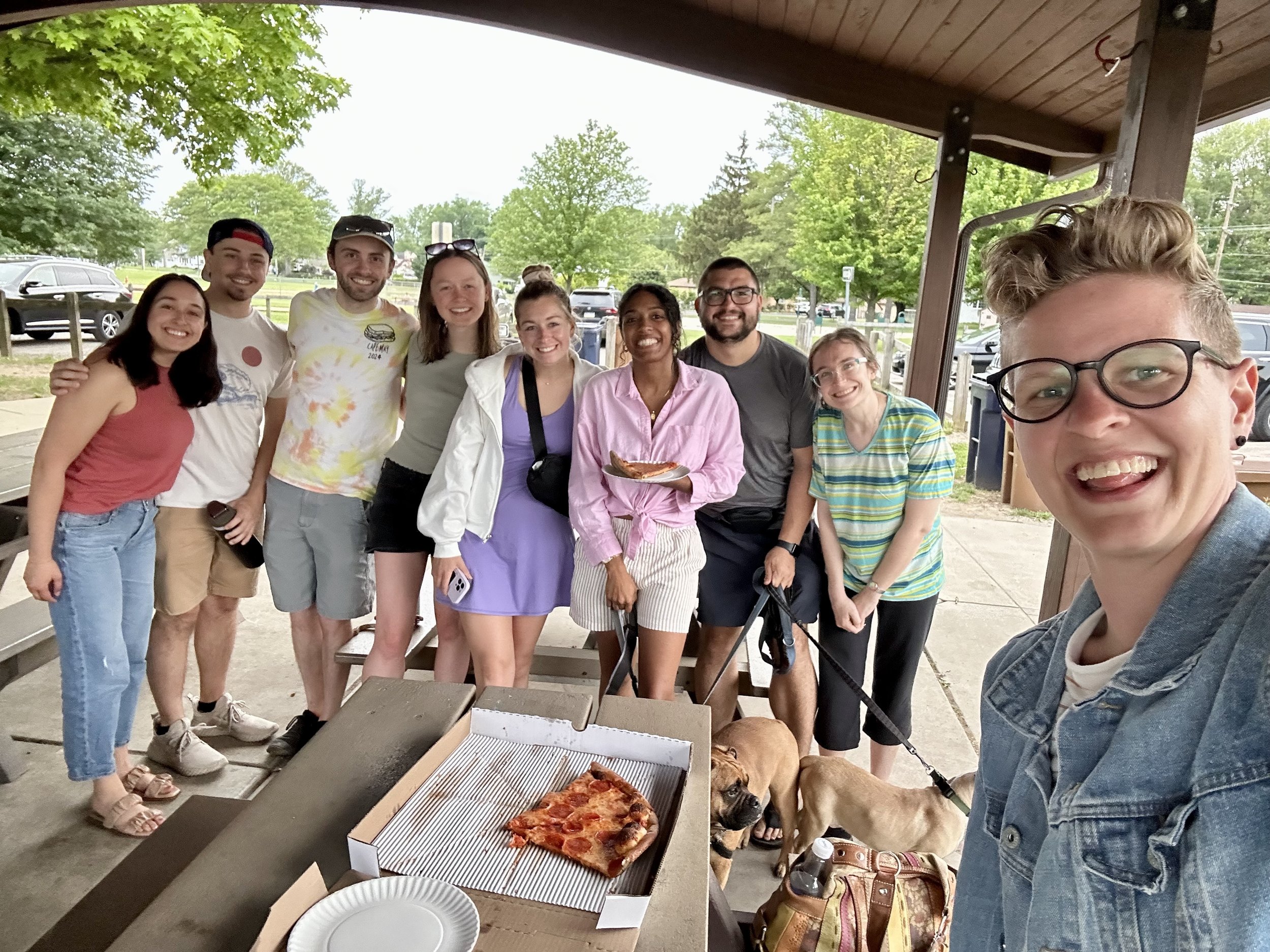 Group of nine young adults standing under a pavilion, smiling, with two dogs and a pizza box in front of them, at an outdoor gathering on a cloudy day.