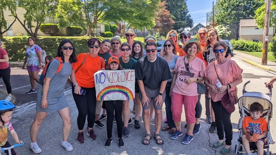 Colorful t-shirts with names and dates displayed outside, with a man smiling among them. A sign reads 'Montco Gun Homicides Past 5 Years'.