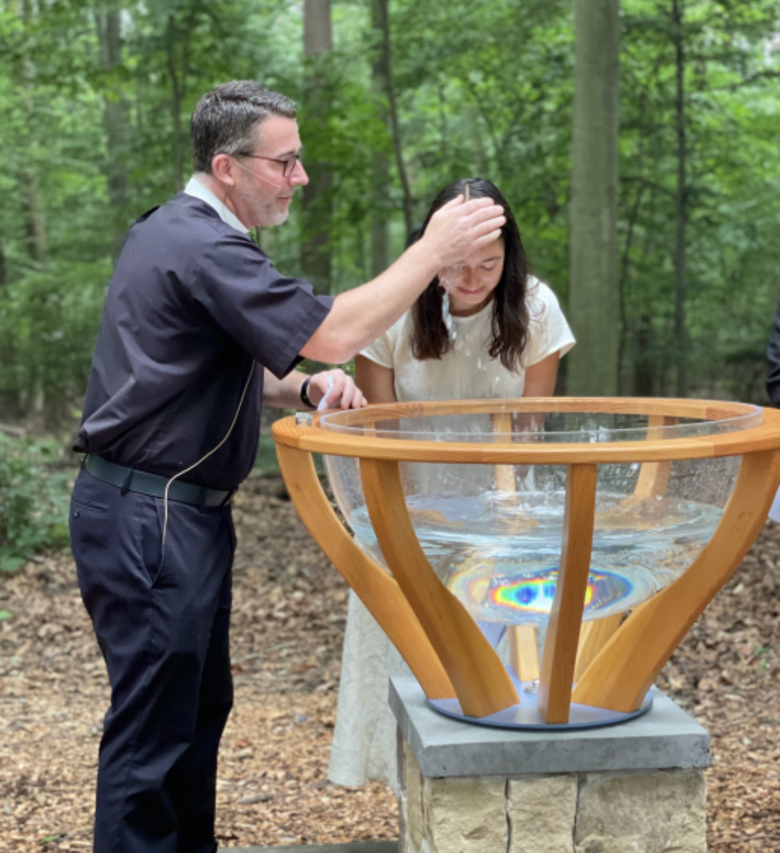A pastor baptizing a woman outdoors in a forested area, with water overflowing in a baptismal font.