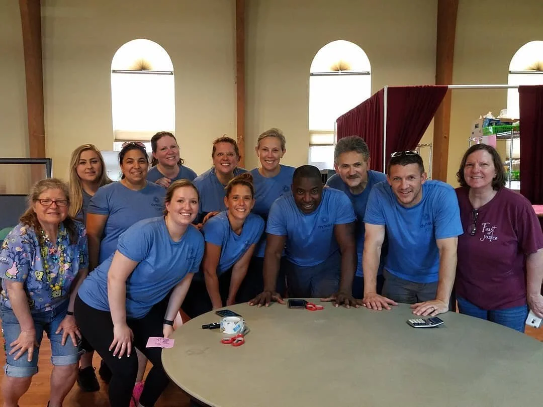 Group of people, mostly wearing blue shirts, gathered around a table in a room with high arched windows and wooden beams, smiling at the camera.