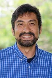 Portrait of a man with dark hair, beard, and mustache, smiling, wearing a blue checkered shirt, with a blurred green outdoor background.