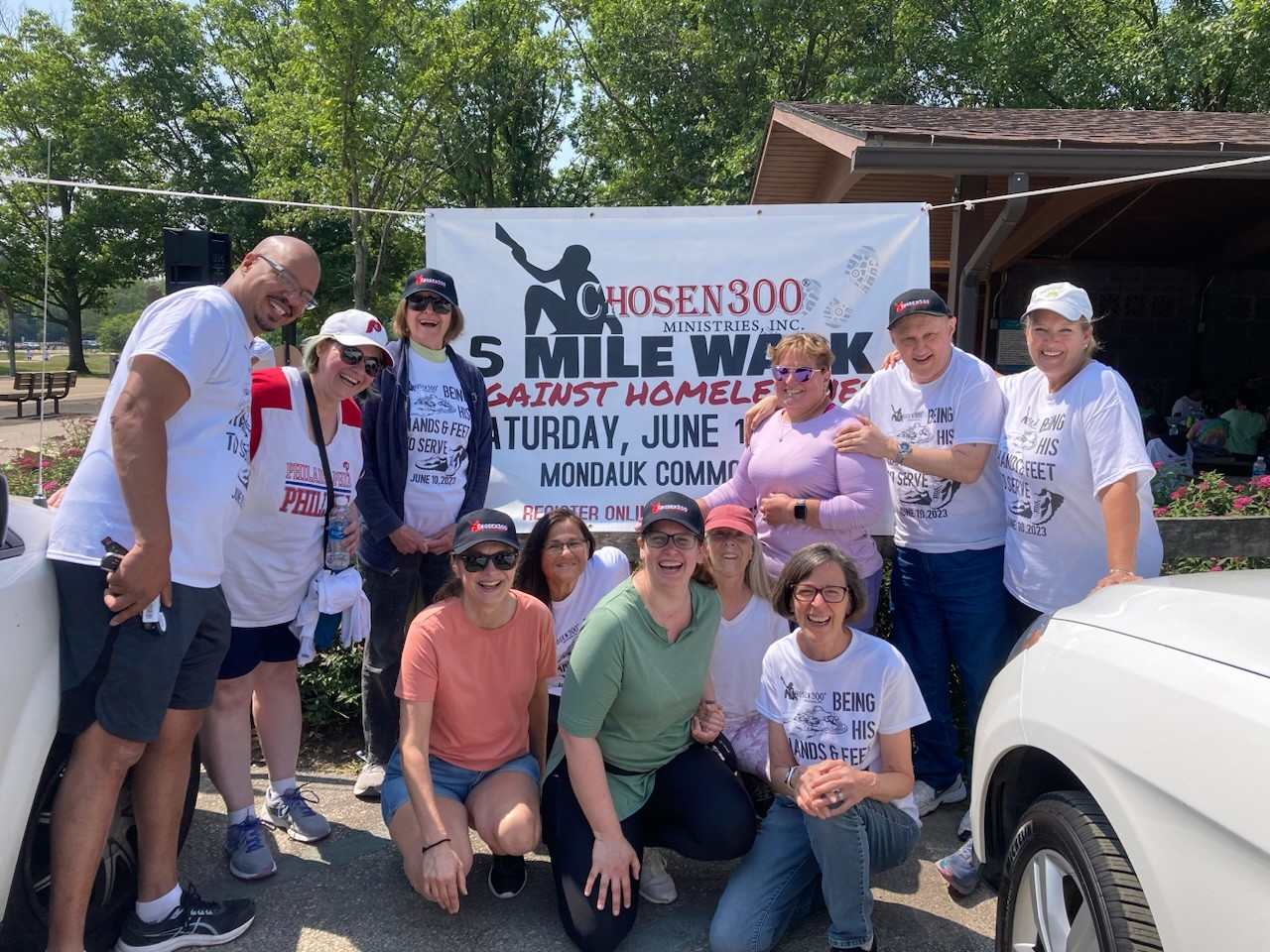 Group of people posing outdoors in front of a banner at a community walk event, with trees and a building in the background.