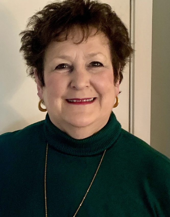 A smiling older woman with short, dark, curly hair, wearing a green turtleneck sweater, gold hoop earrings, and a gold necklace, standing in front of a plain background.