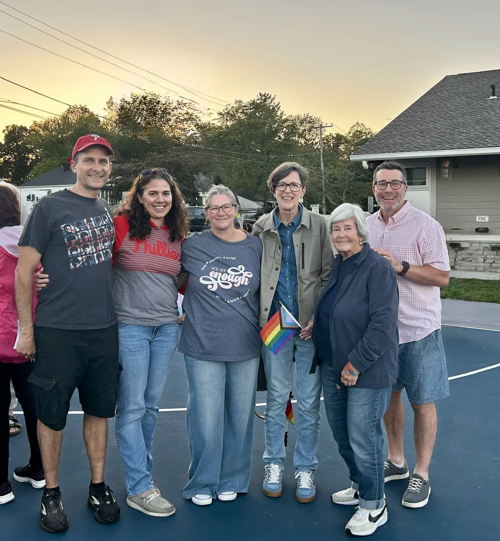 A diverse group of seven people standing outside on a sports court during sunset, holding rainbow pride flags, smiling at the camera.