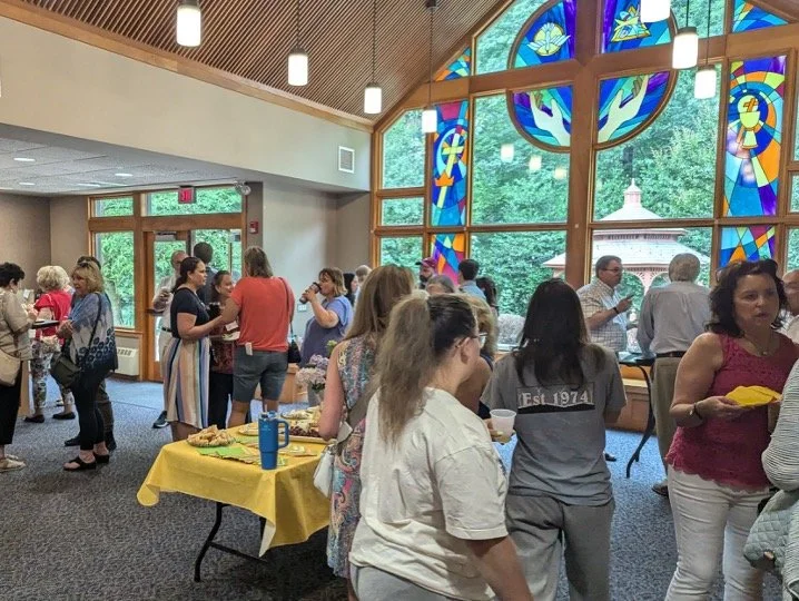 A group of people gathered indoors around tables with food and drinks during a social event or reception in a room with large stained glass windows and wooden beams.