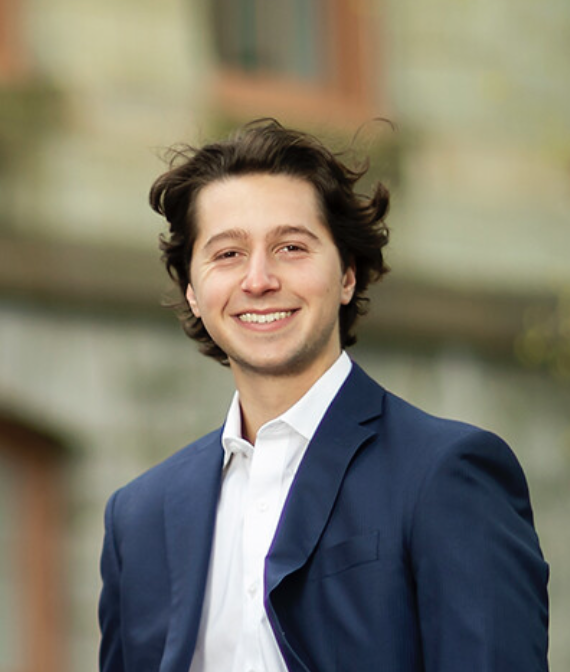 Young man with dark, wavy hair wearing a navy suit and white shirt, smiling outdoors with a blurred building background.