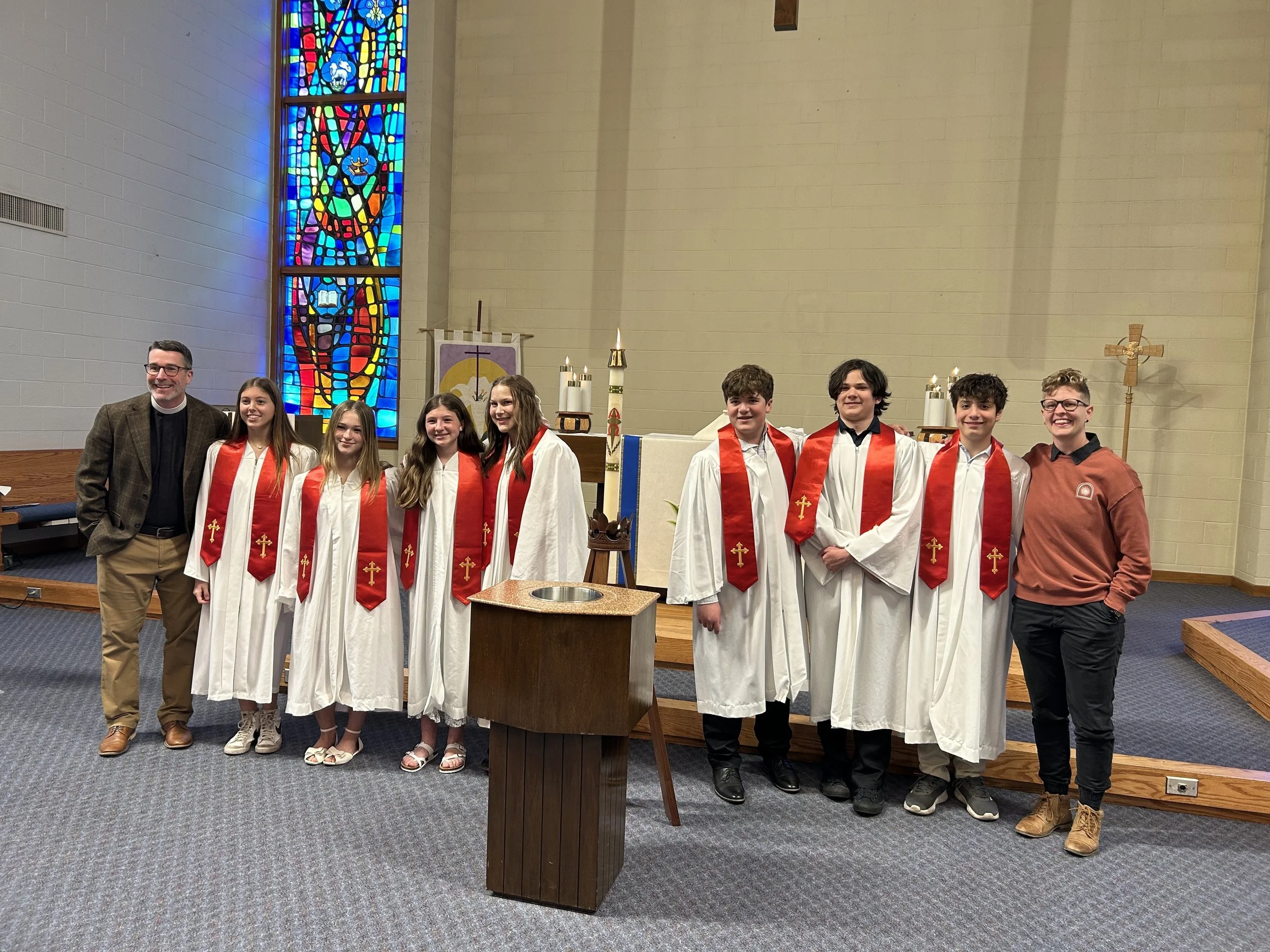Group of nine people, including six boys and two girls in white robes with red stoles, standing in church sanctuary with a priest and a woman, near altar with candles, cross, and stained glass window.