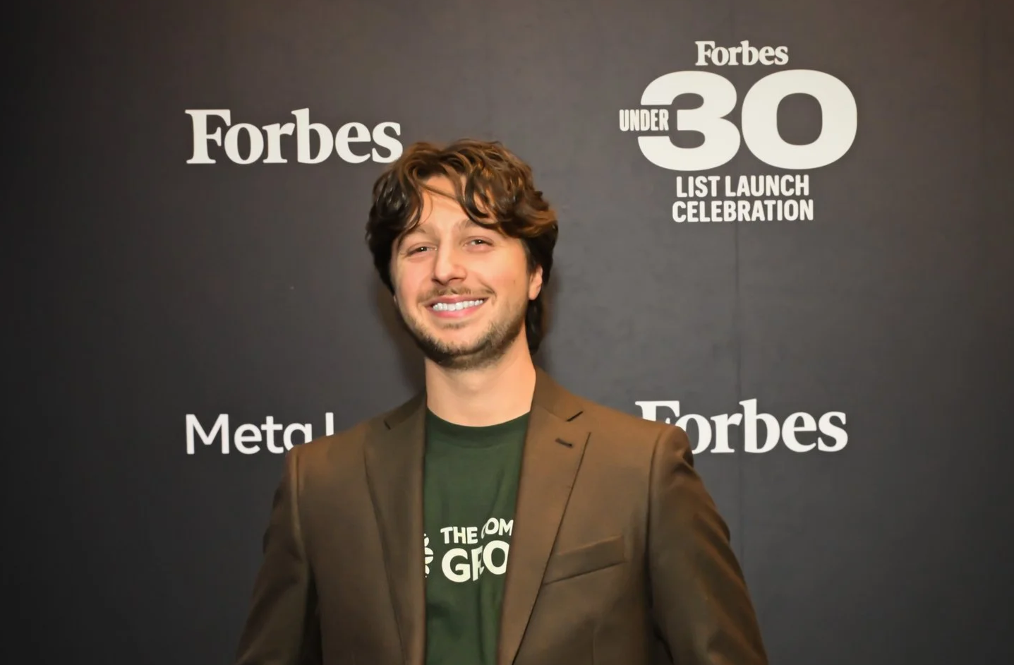 A man with brown hair and a beard, wearing a brown blazer and a green t-shirt, smiling in front of a black backdrop with white text reading 'Forbes 30 Under 30 List Launch Celebration'.