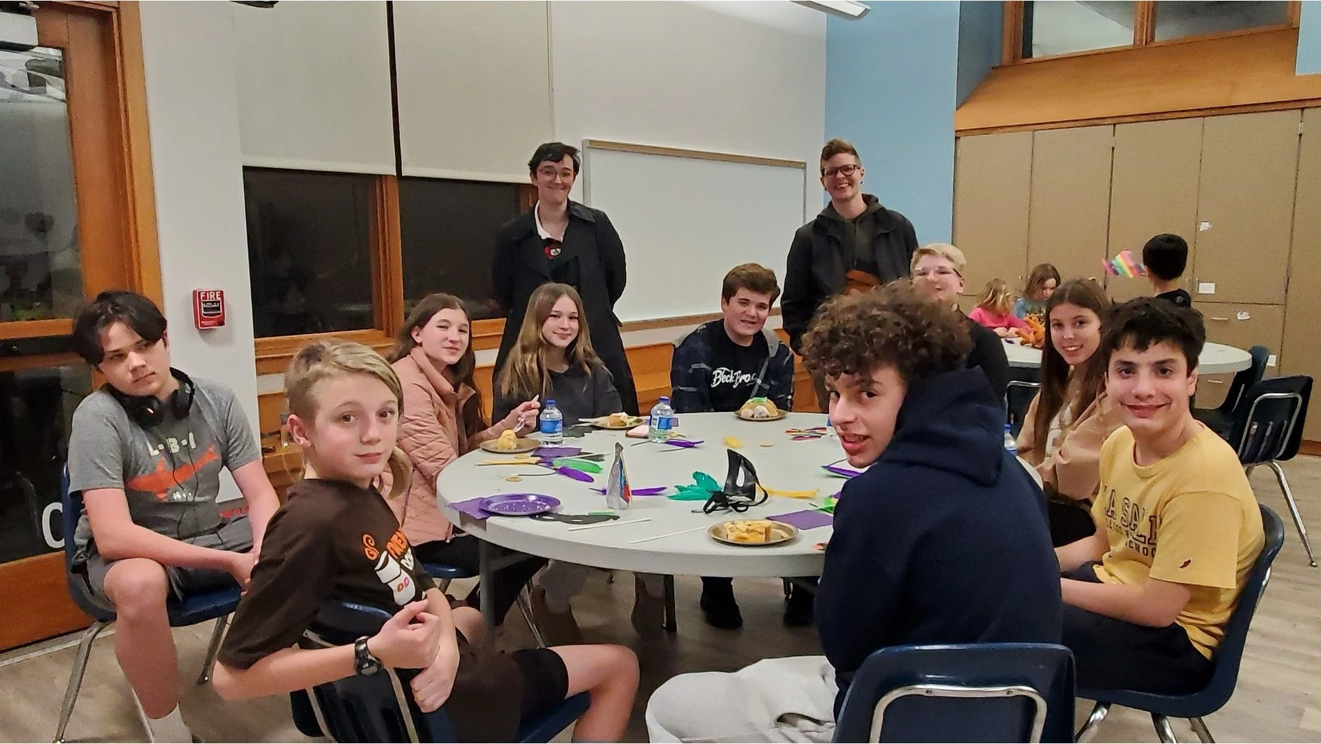 Group of children and teenagers gathered around a circular table in a room, with two adults standing behind them, some of the children have plates of food, and colorful paper decorations are on the table.