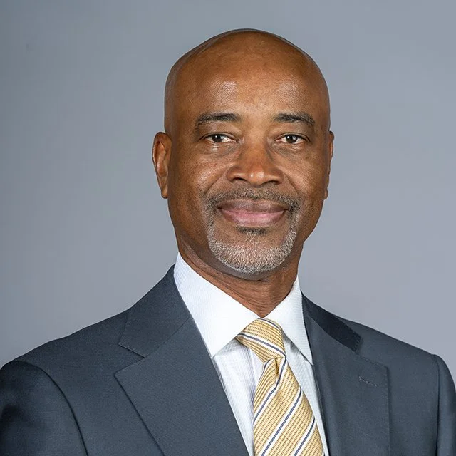 A middle-aged African American man with a bald head and gray beard, wearing a dark suit, white shirt, and yellow striped tie, smiling against a gray background.