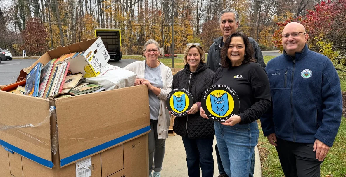 Group of five people standing outdoors near a large box filled with books, some holding circular signs that say 'Read it. Love it. Donate it. #booksmiles.org.' The background includes trees with autumn foliage and a parking lot.