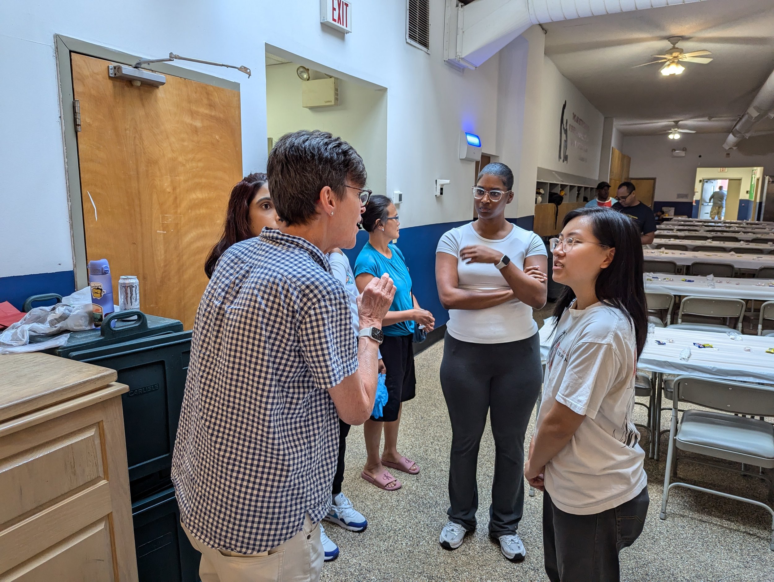 Group of five people engaged in conversation inside a community center or cafeteria, with tables and chairs in the background.