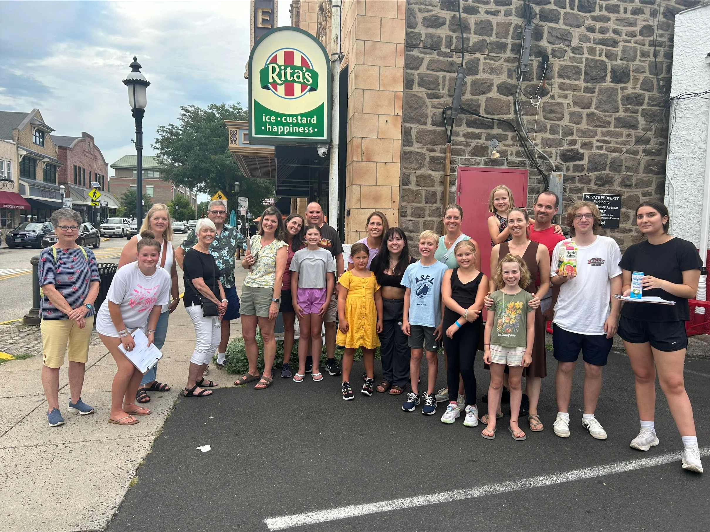 A group of adults and children standing on a sidewalk in front of a bakery called Rita's Ice Custard Happiness.