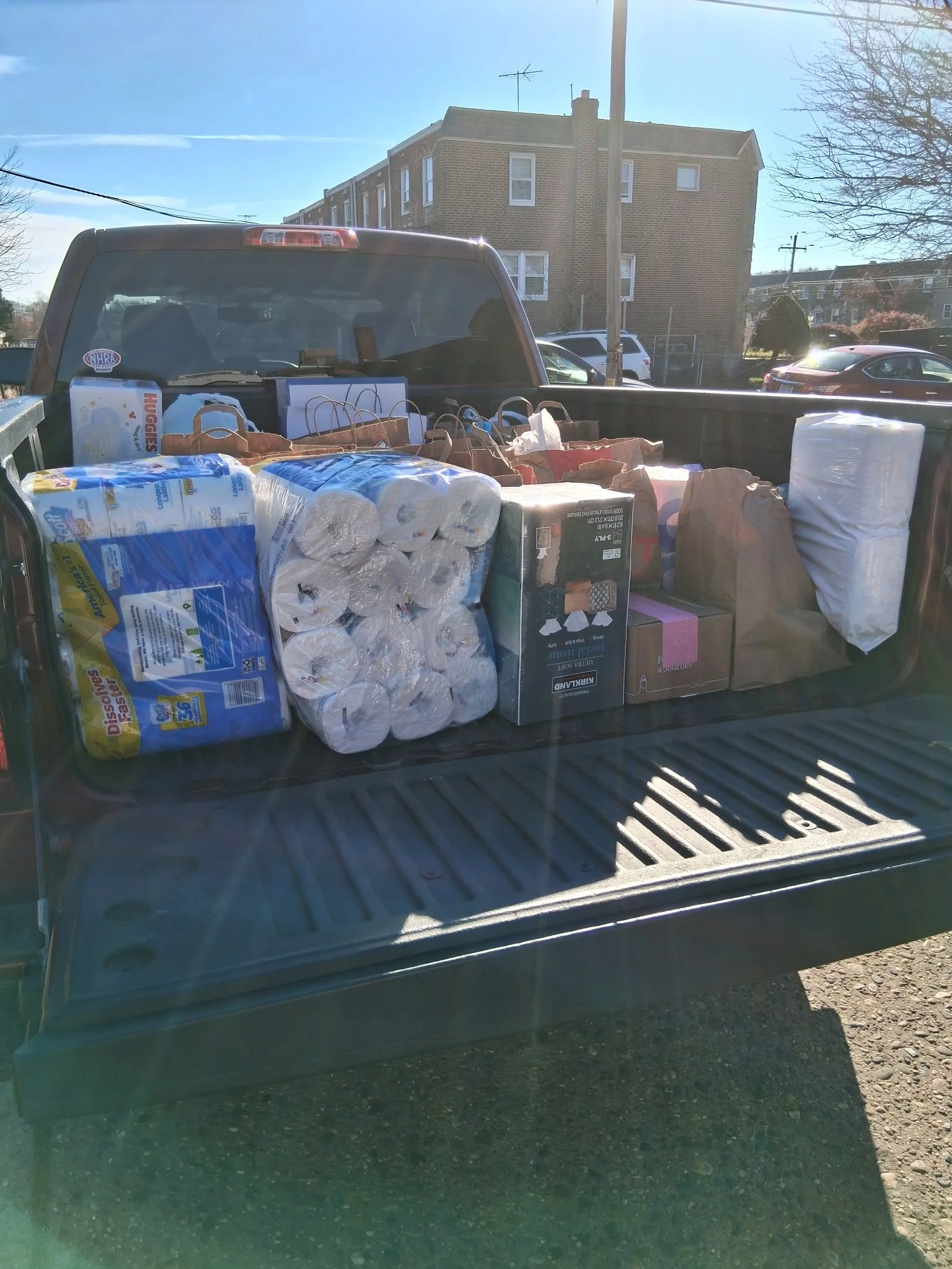 The back of a truck loaded with paper towels, toilet paper, paper bags, and other household items parked in a residential area with buildings and parked cars in the background.