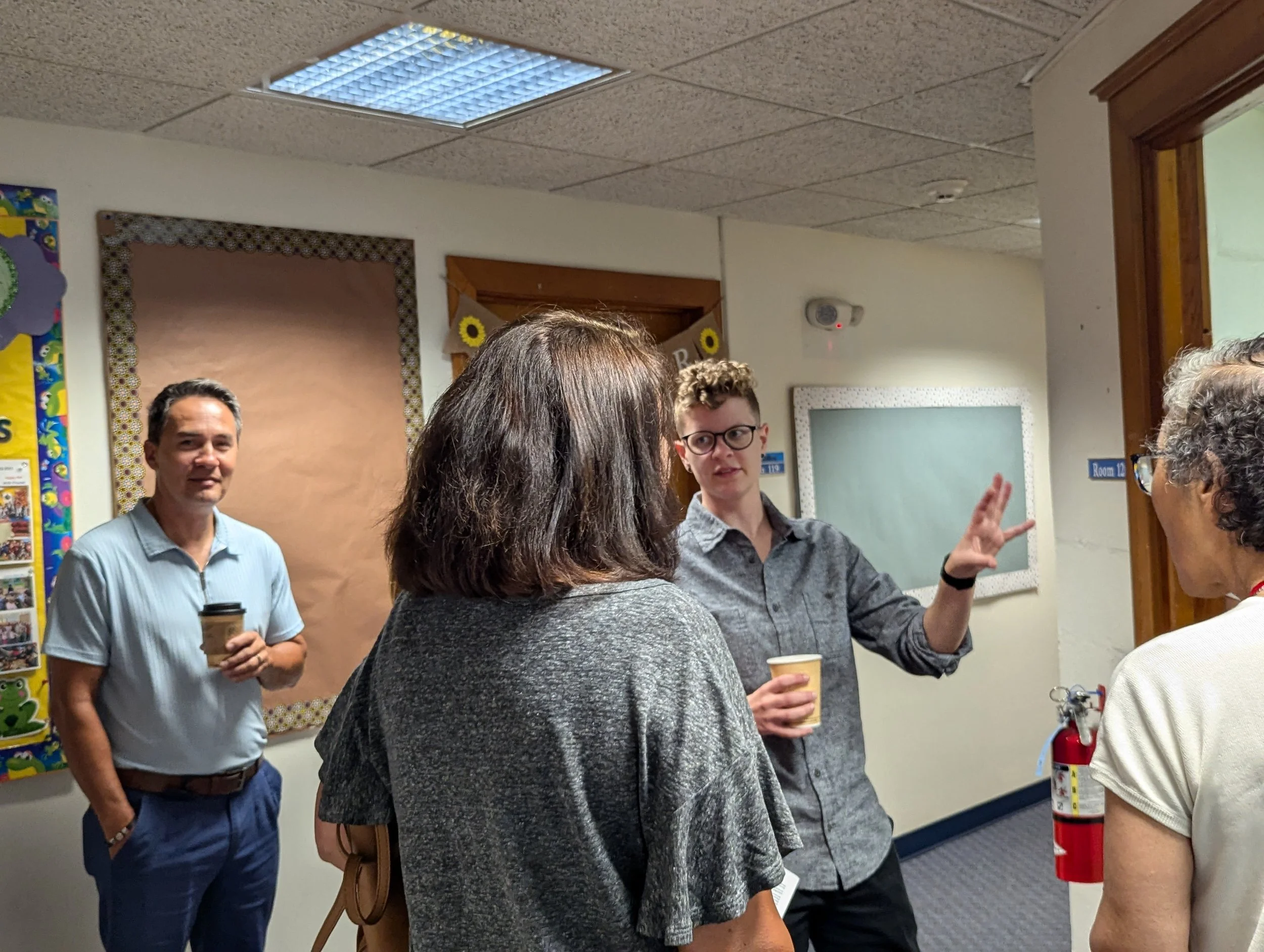 Group of people engaged in conversation in a classroom or office setting, with a bulletin board, a fire extinguisher, and a door in the background.
