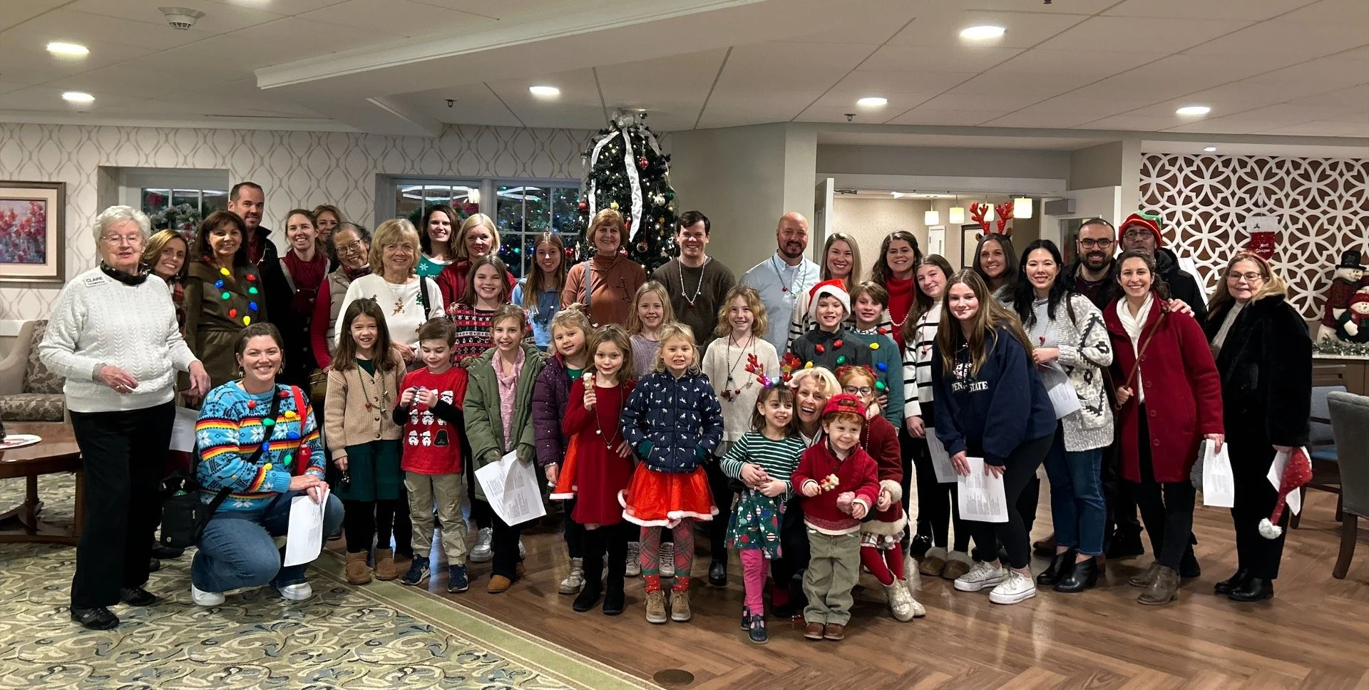 Large multigenerational group in festive Christmas sweaters posing around a decorated Christmas tree indoors