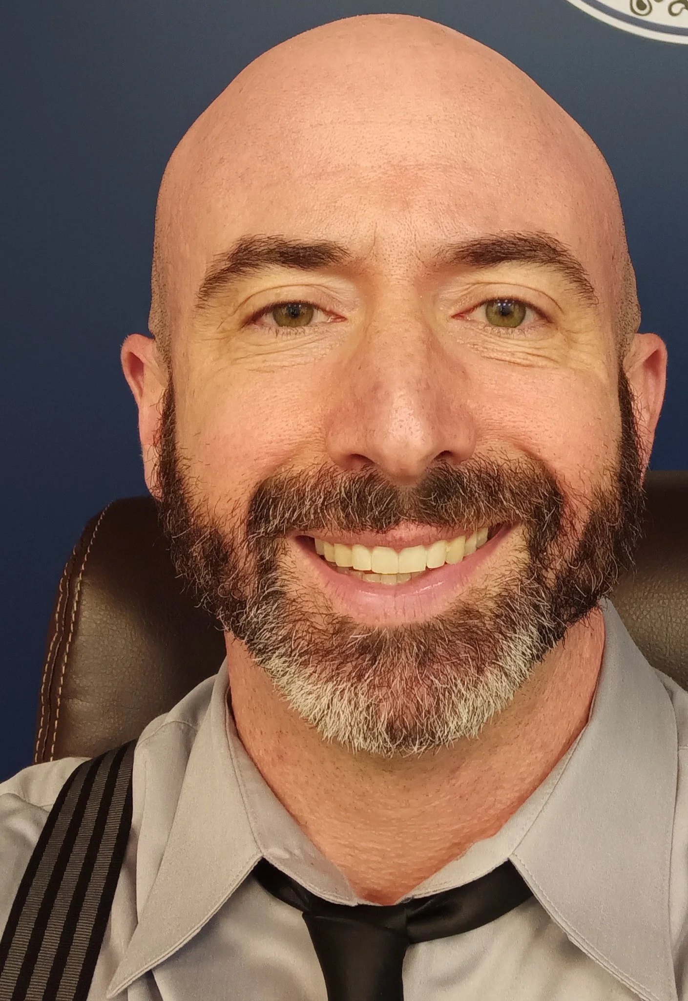 A close-up photo of a smiling man with a bald head, a full beard, wearing a gray uniform with black suspenders and a black tie, seated on a chair with a blue background.