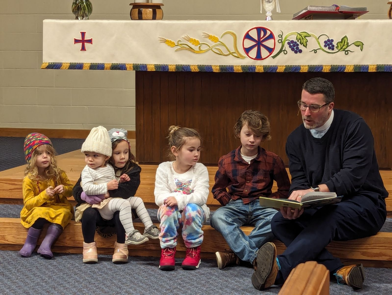 A group of six children sitting on wooden steps in a church, with a priest or pastor reading from a book. The children are dressed in colorful clothing and winter hats. The church interior has a decorated altar with a white cloth, a cross, and art on it.