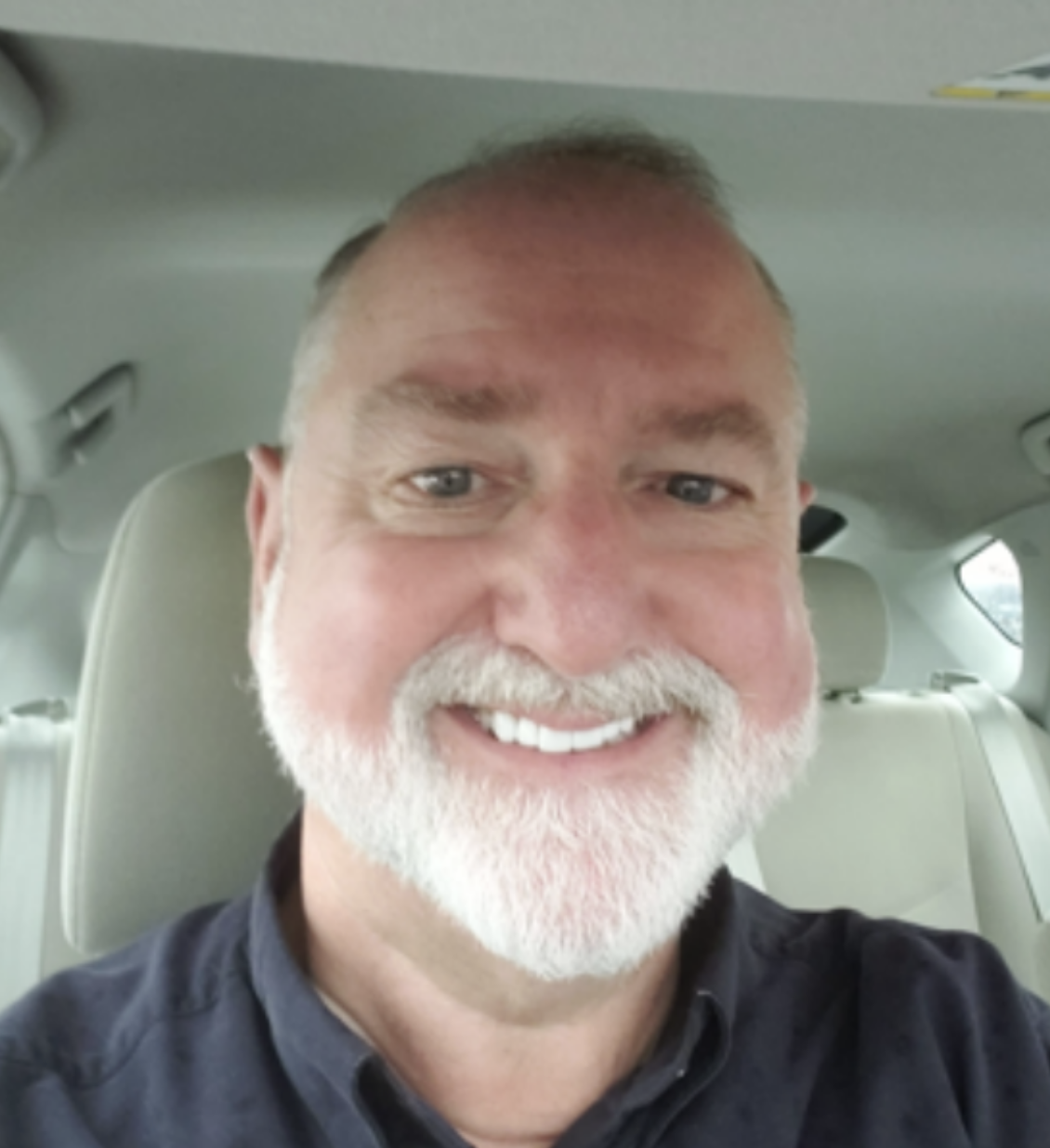 A smiling man with a white beard and short hair sitting in the backseat of a car.
