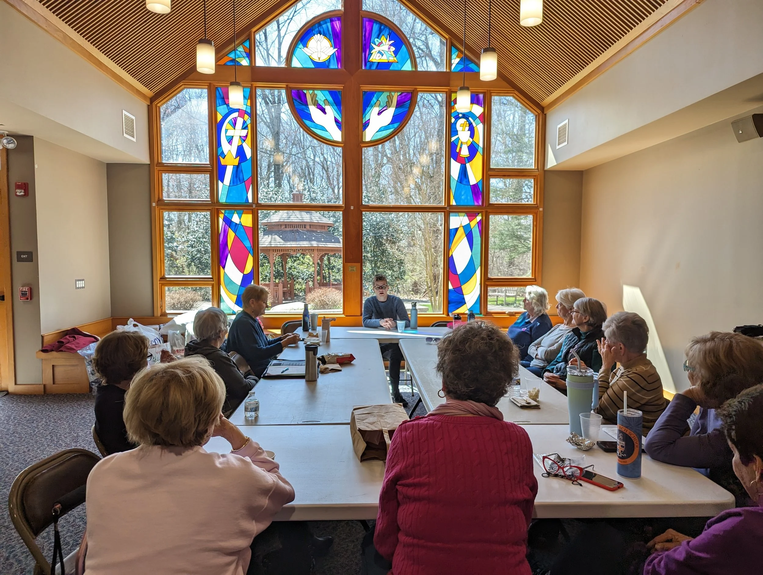 Group of women seated around a U-shaped table for a meeting inside a room with large stained glass windows depicting religious symbols and figures, and an outside view of trees and a gazebo.