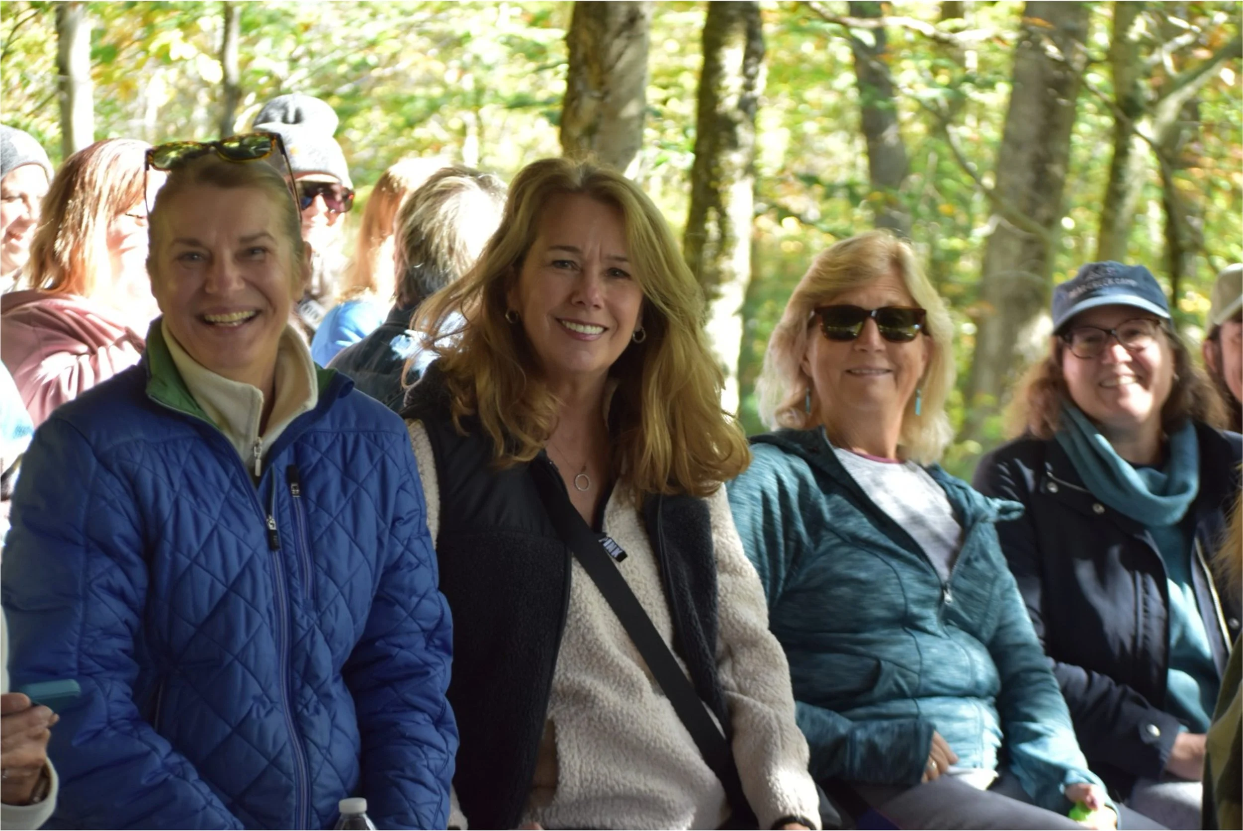 A group of women smiling outdoors in a wooded area, dressed in casual and outdoor clothing.