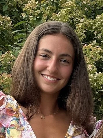 Smiling young woman with long brown hair posing outdoors in front of flowering greenery