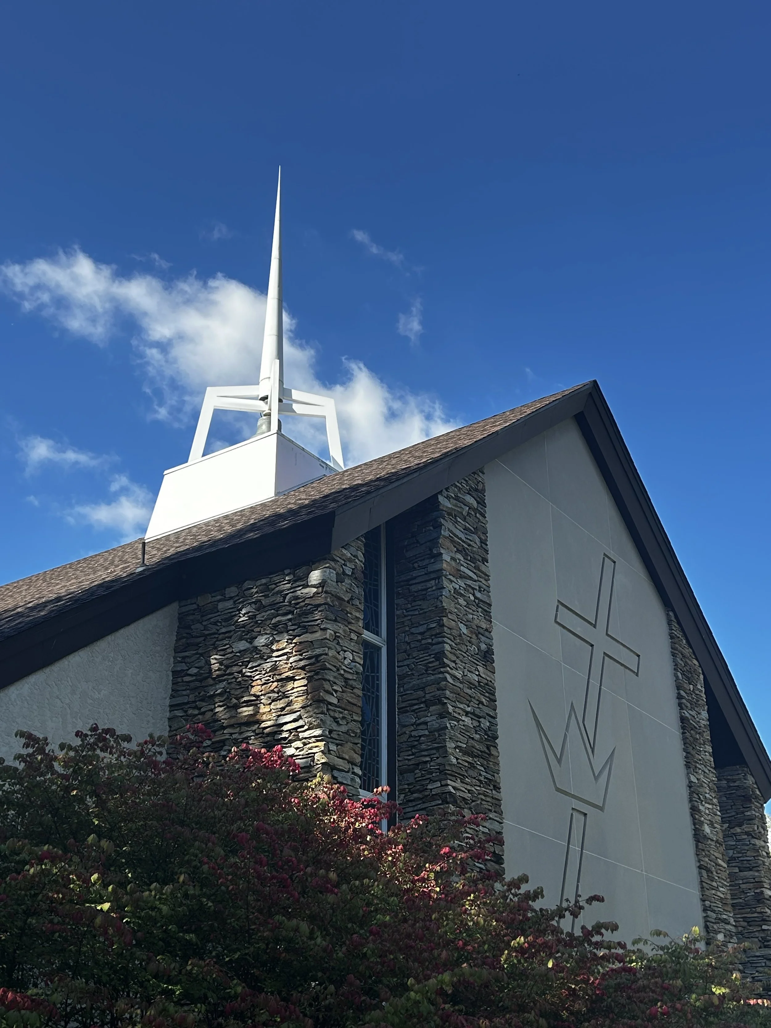 A group of children and adults sitting on a wooden platform during a church gathering. A man in religious attire is speaking to the children, with some children showing curiosity and engagement. The background has a colorful stained glass window and religious banners.