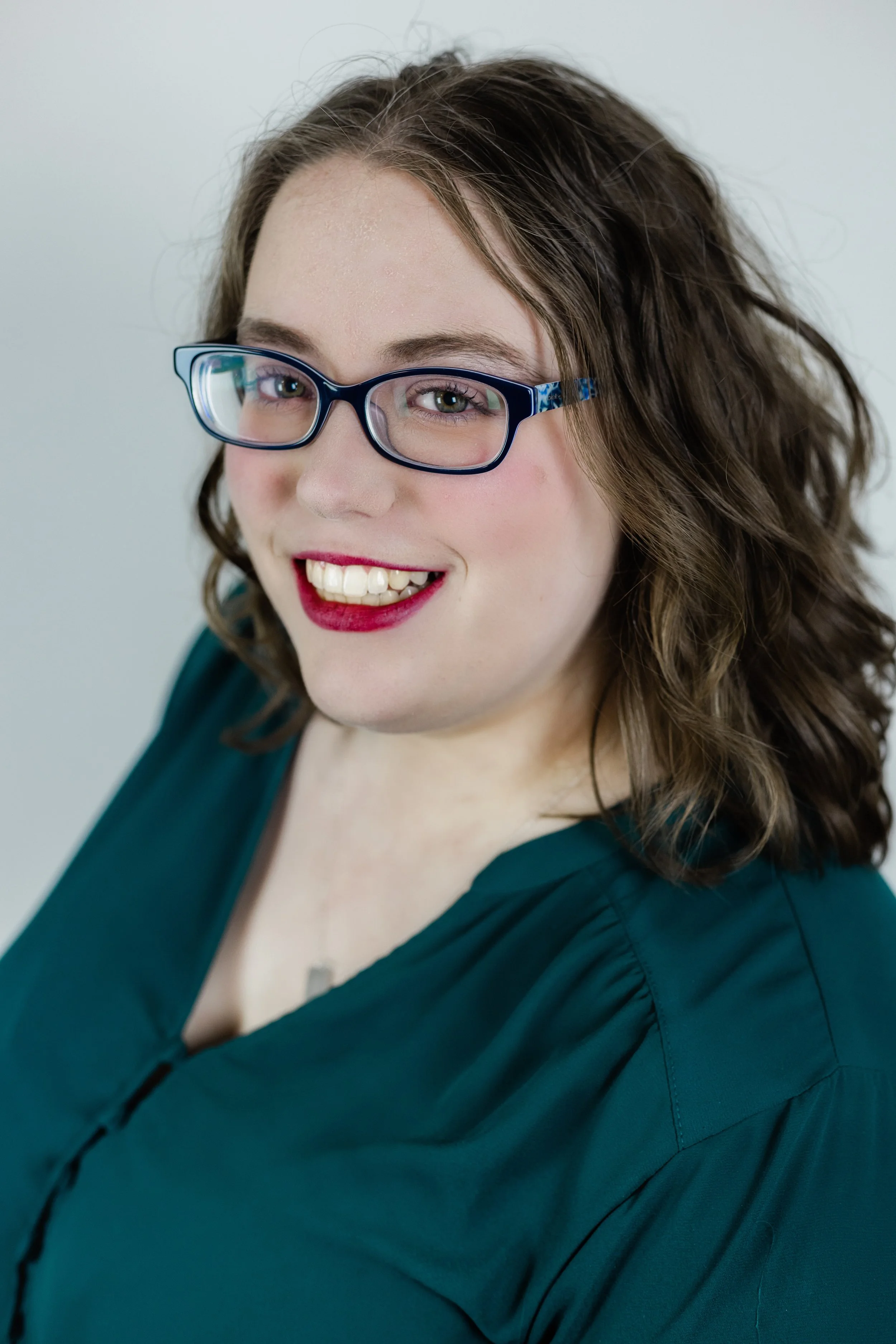 A woman with curly brown hair, glasses, and red lipstick smiling for the camera against a plain light gray background.