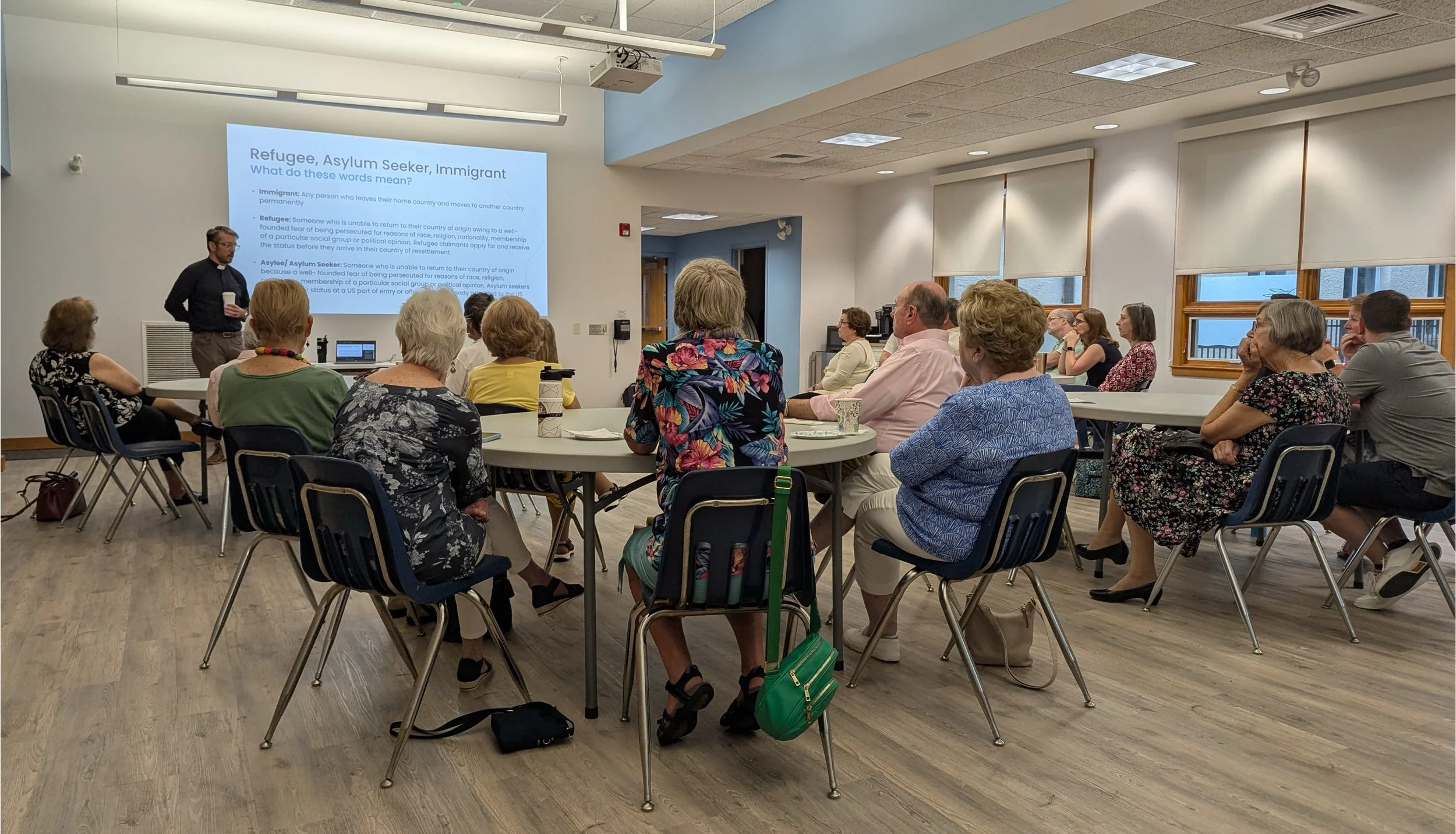 A group of people attending a classroom lecture or presentation. They are seated at round tables, listening to a speaker standing next to a projector screen displaying information about refugees, asylum seekers, and immigrants. The room has windows letting in natural light.