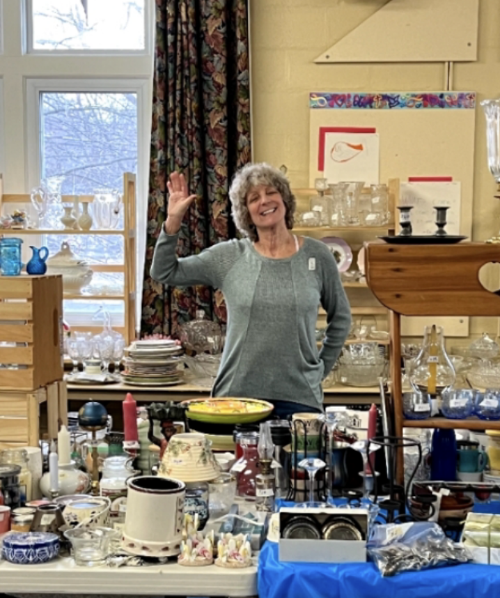 Smiling woman waving at a table filled with various glassware, dishes, candles, and decorative items in an indoor setting with a window and patterned curtains in the background.